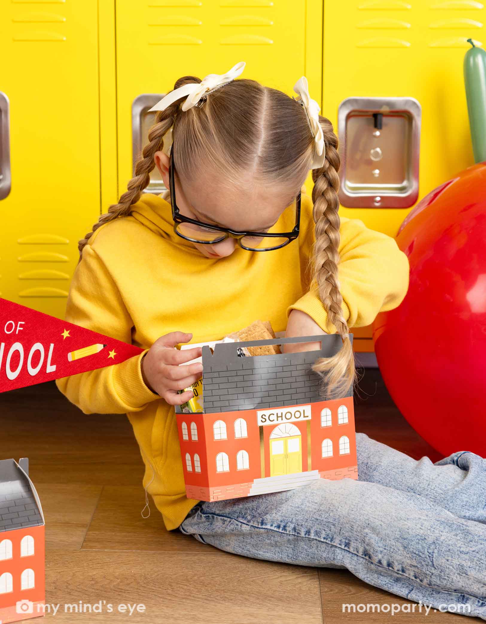 A girl in her yellow sweatshirt and glasses looking into Momopartys's school building shaped treat box. She is sitting in front of a first day of school celebration decoration featuring various school themed party decors from Momopartys including a jumbo apple latex balloon, first day of preschool party pennant, and school shaped treat boxes.
