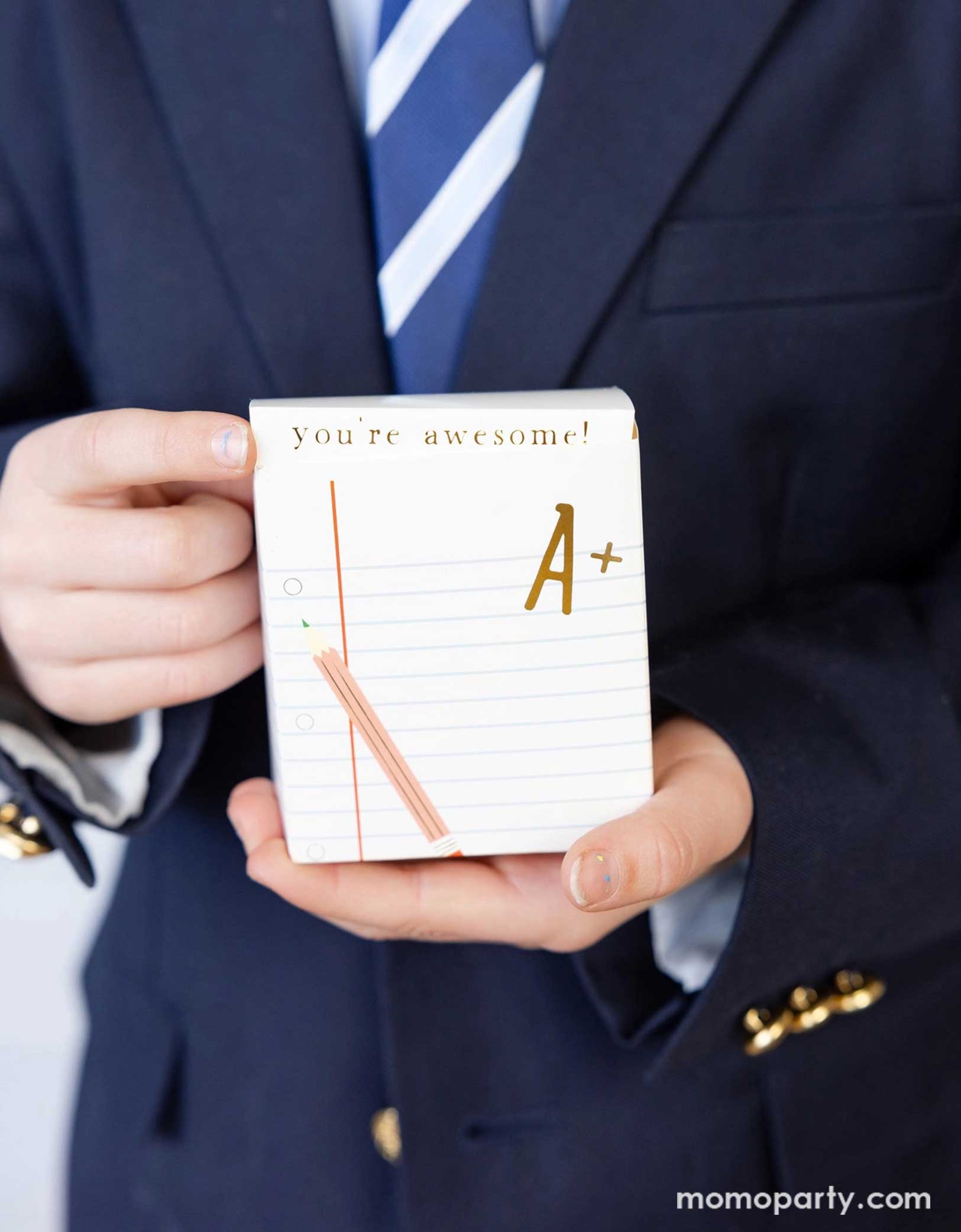 A boy in his school uniform holding Momopartys's 3" x 2" x 5.5" notebook treat box. Cleverly disguised as notebooks with "You're awesome!" on it, this fun container add a scholarly twist to your back-to-school festivities. Perfect for surprising students with sweet goodies while keeping the academic theme alive!
