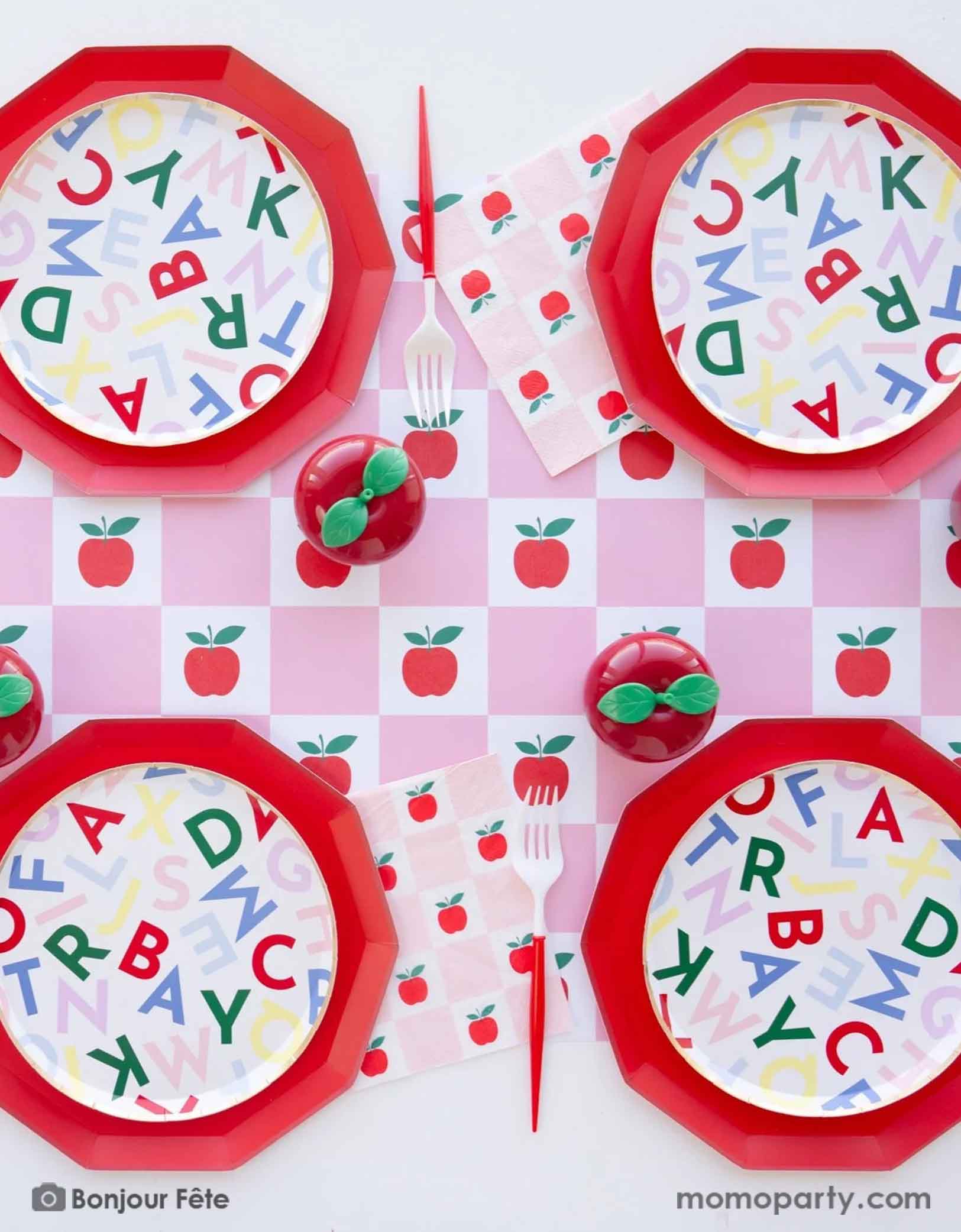 A festive back to school apple themed party table setting featuring various apple and school themed party tableware from Momopartys including large red dinner plates, alphabet side plates, red and white utensils, apple and pink checkered table runner and matching large napkins. With some cute apple shaped centerpiece in the middle, this makes a perfect tablescape for kid's back to school party or first day of school celebration.
