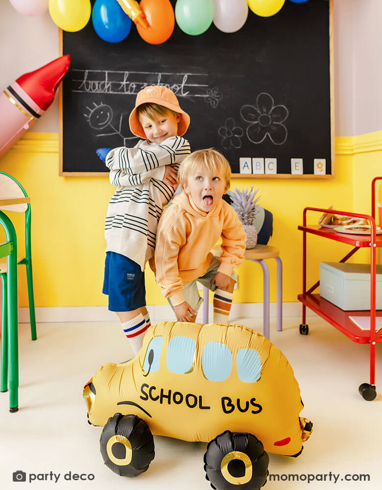 Two school aged boys making funny faces in a school classroom setting decorated with Momopartys's back to school party balloons including a school bus shaped foil balloon, a red crayon shaped foil balloon, a few festive colorful latex balloons hung above the chalkboard. With colorful, vibrant school furniture around it, this makes a great inspo for a modern and playful back to school, first day of school celebration in a school set up.