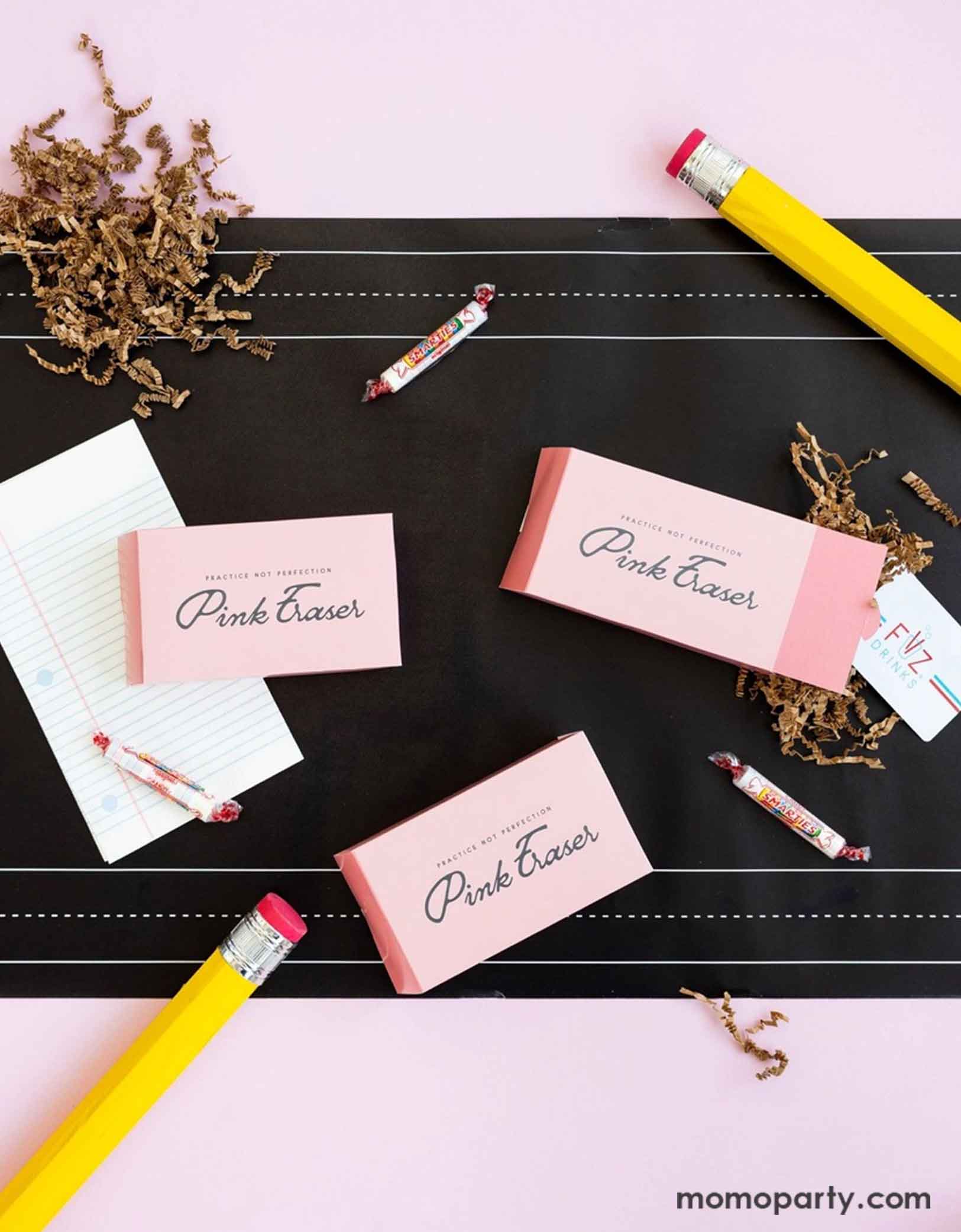 An overhead shot of a back to school party table featuring various school themed party supplies from Momopartys including pink eraser shaped treat boxes, notebook shaped napkins, and chalk board paper table runner. Around the party supplies are some smarty candies, a couple of jumbo pencils as props -  this table settings is a perfect inspiration for a simple yet adorable idea for kid's goodie bags at a school themed event or celebration.