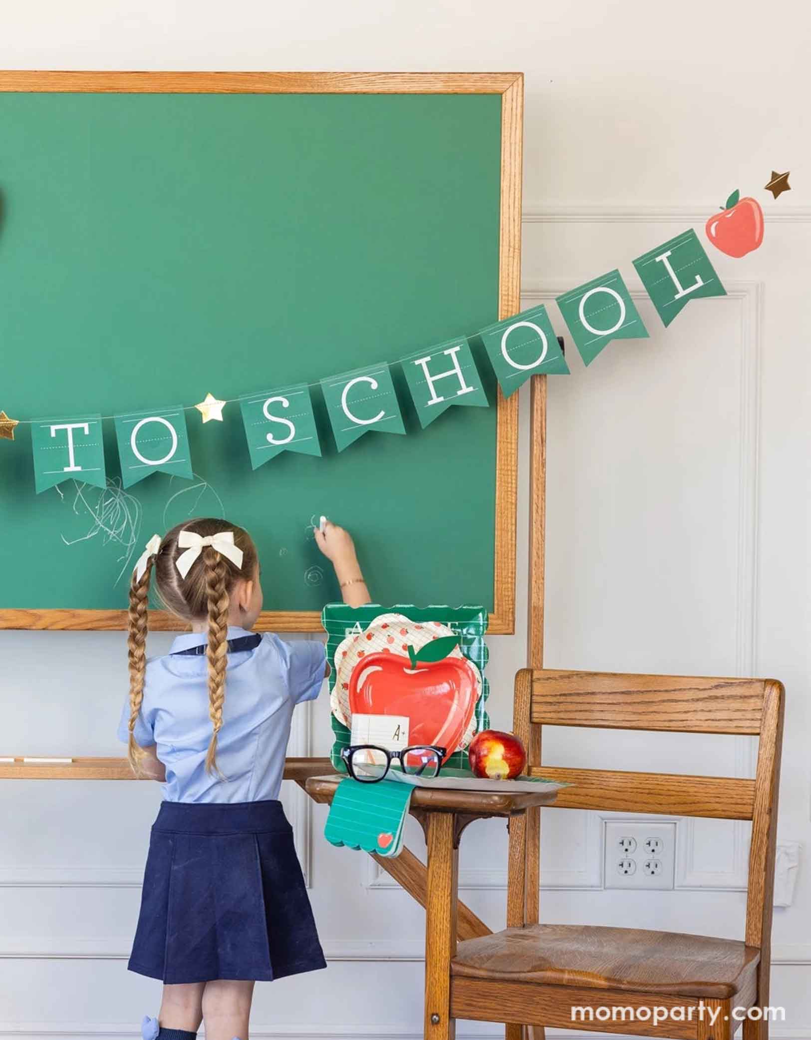 A school-aged girl in her blue uniform writing something on a chalkboard in a classroom set up decorated with a bunch of school themed party supplies from Momopartys including 8.5" apple shaped plates, 10" apple pattern plates, alphabet dinner plates and matching napkins. The chalkboard is adorned with the back to school party banner hung across, with an apple shaped plate on it - an adorable and cute set up to celebrate kid's back to school season!