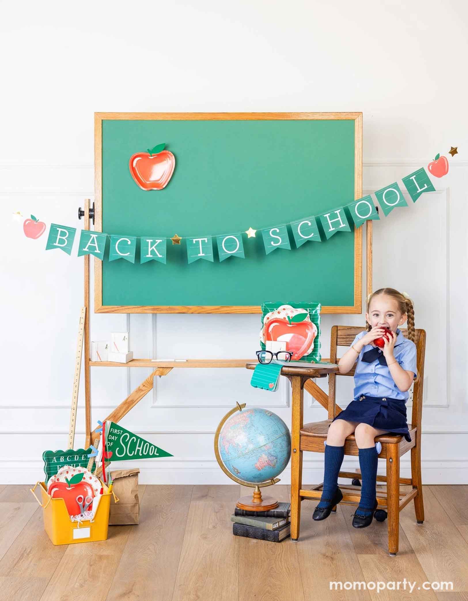 A school-aged girl in her blue uniform eating an apple on a school chair in a classroom setting decorated with a bunch of school themed party supplies from Momopartys including 8.5" apple shaped plates, 10" apple pattern plates, alphabet dinner plates and matching napkins. Behind her is a classroom chalkboard with back to school party banner hung across with an apple shaped plate on it - an adorable and cute set up to celebrate kid's back to school season!