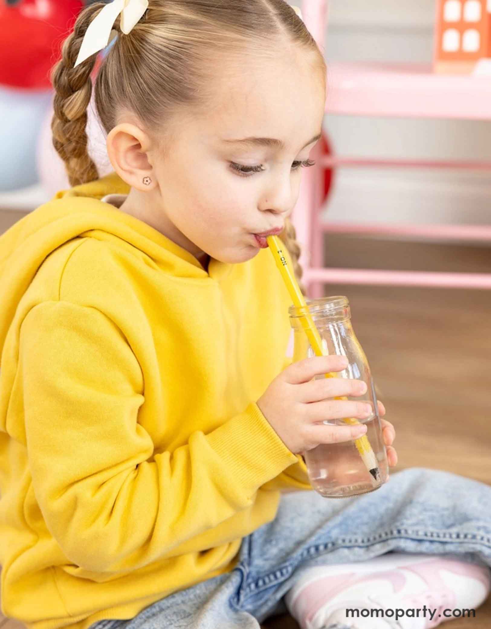 A little girl wearing a yellow sweatshirt sipping water with Momopartys's pencil shaped straw from a milk bottle. This eco-friendly reusable straw is perfect for your party and come in a fun pencil shape. It's perfect for kid's back to school party or first day of school celebration.