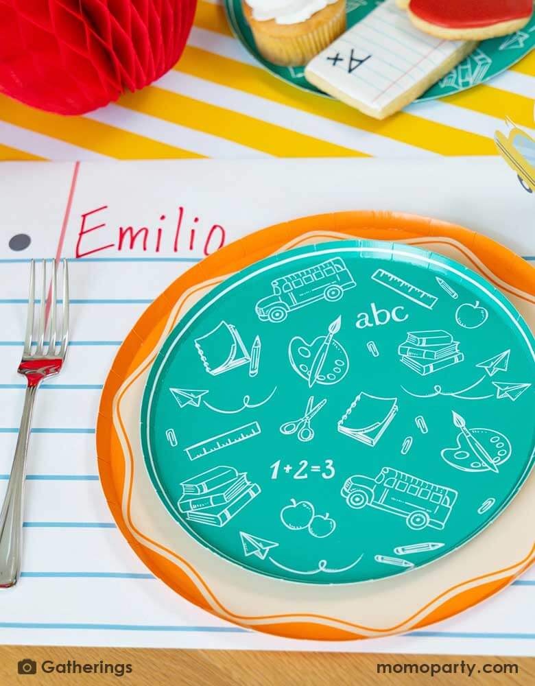 A festive back to school first day of school celebration party table place setting featuring Momopartys's back to school themed party supplies including the school day assorted side plates with school supplies designs in green, and the loose leaf paper placemat by Gatherings. With school supplies shaped sugar cookies around and a honeycomb apple decoration on the table, makes a great inspo to celebrate the start of the new school year with kids!
