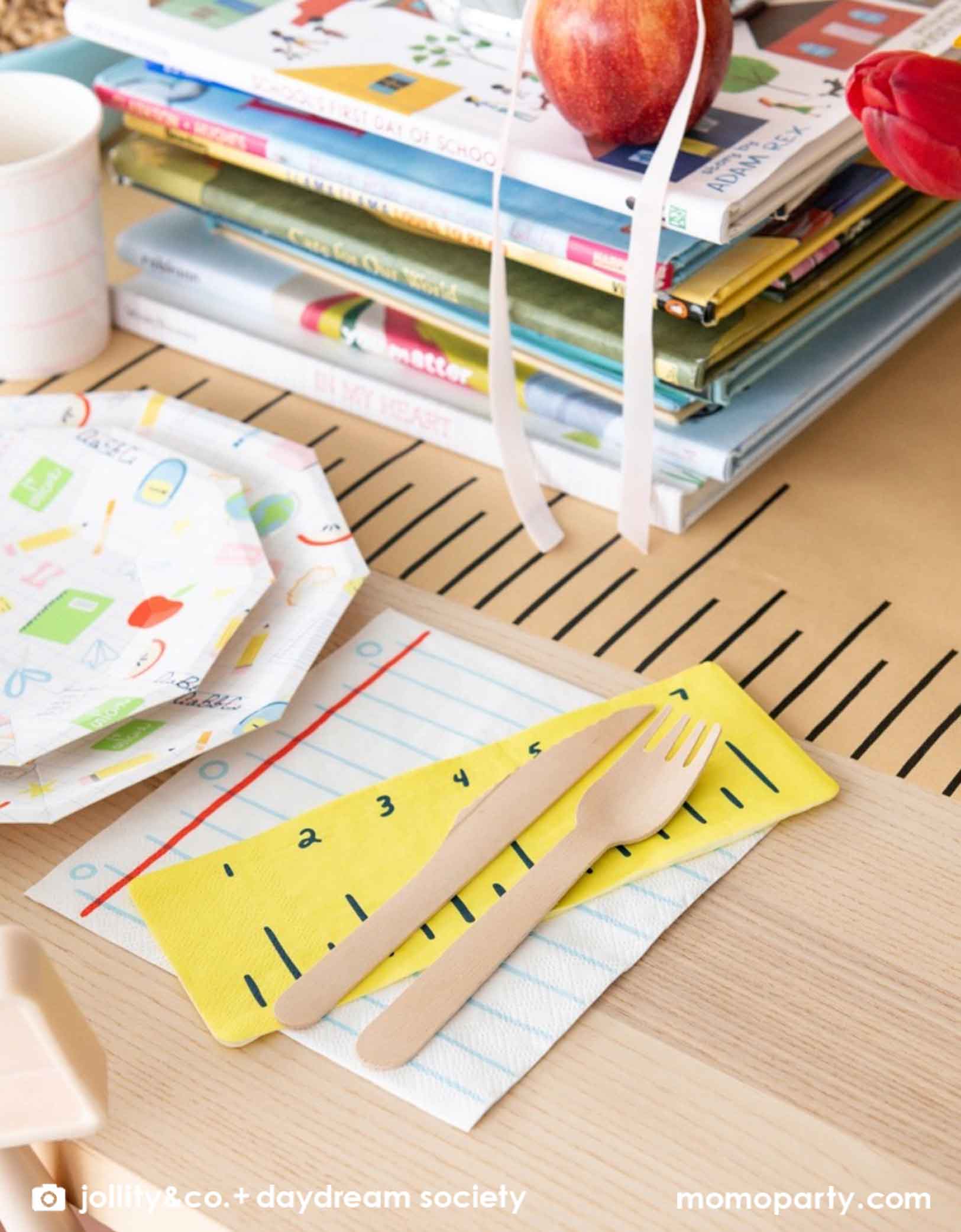 A close up shot of an adorable kid's school themed party table featuring school supplies themed party tablewares from Momopartys including the Smarty Pants ruler shaped napkins and notebook shaped napkins along with large and small school icons patterned plates, all on a ruler inspired table runner. In the middle of the table there is a pile of books and some apples as centerpiece, making this a great decoration inspiration for kid's  first day of school celebration or a back to school party.