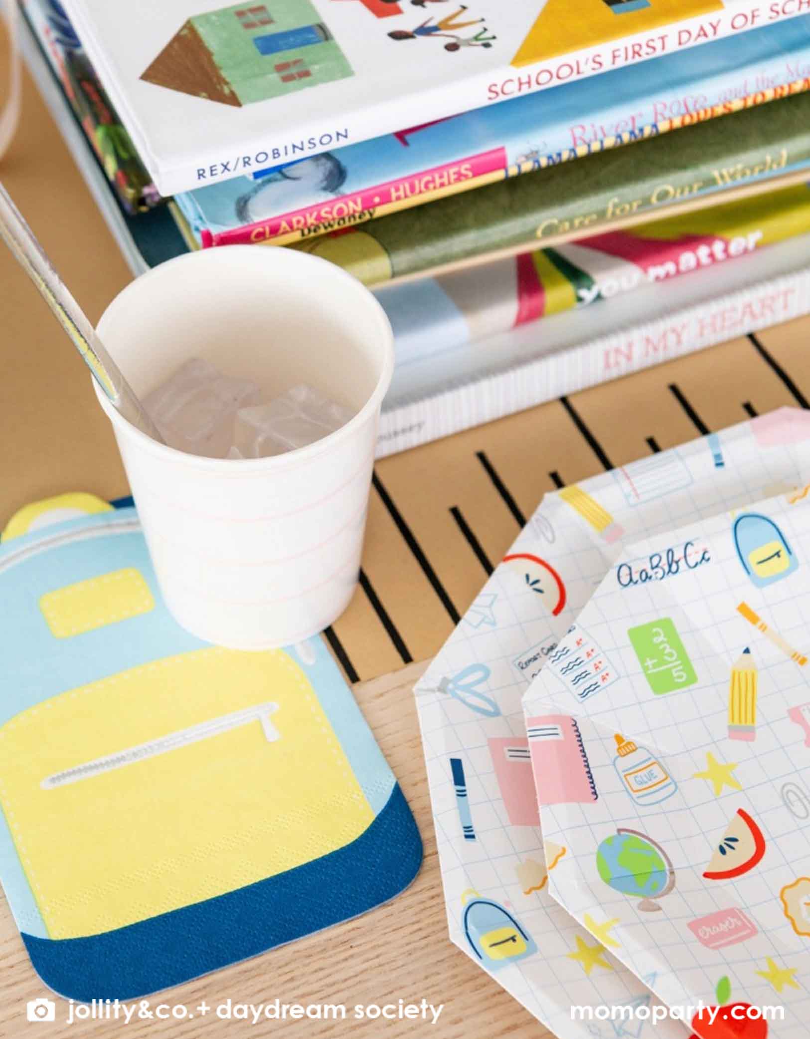 A close up shot of an adorable kid's school themed party table featuring school supplies themed party tablewares from Momopartys including the Smarty Pants backpack shaped napkin, a pink striped party cup, along with large and small school icons patterned plates, all on a ruler inspired table runner. In the middle of the table there is a pile of books as centerpiece, making this a great decoration inspiration for kid's  first day of school celebration or a back to school party.