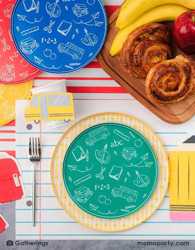 A festive back to school first day of school celebration party table setting featuring Momopartys's back to school themed party supplies including the No. 2 pencil patterned dinner plates, school day side plates with school supplies designs in four different colors, and the notebook inspired placemat by Gatherings. With pencil shaped napkins and red backpack shaped napkins, plus a plate of treats and fruits, this makes a great inspo to celebrate the start of the new school year with kids!