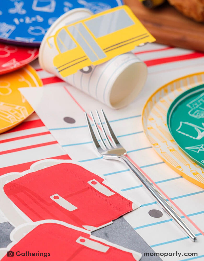 A festive back to school first day of school celebration party table setting featuring Momopartys's back to school themed party supplies including the No. 2 pencil patterned dinner plates, school day side plates with school supplies designs in four different colors, and the notebook inspired placemat by Gatherings. With pencil shaped napkins and red backpack shaped napkins, plus a plate of treats and fruits, this makes a great inspo to celebrate the start of the new school year with kids!