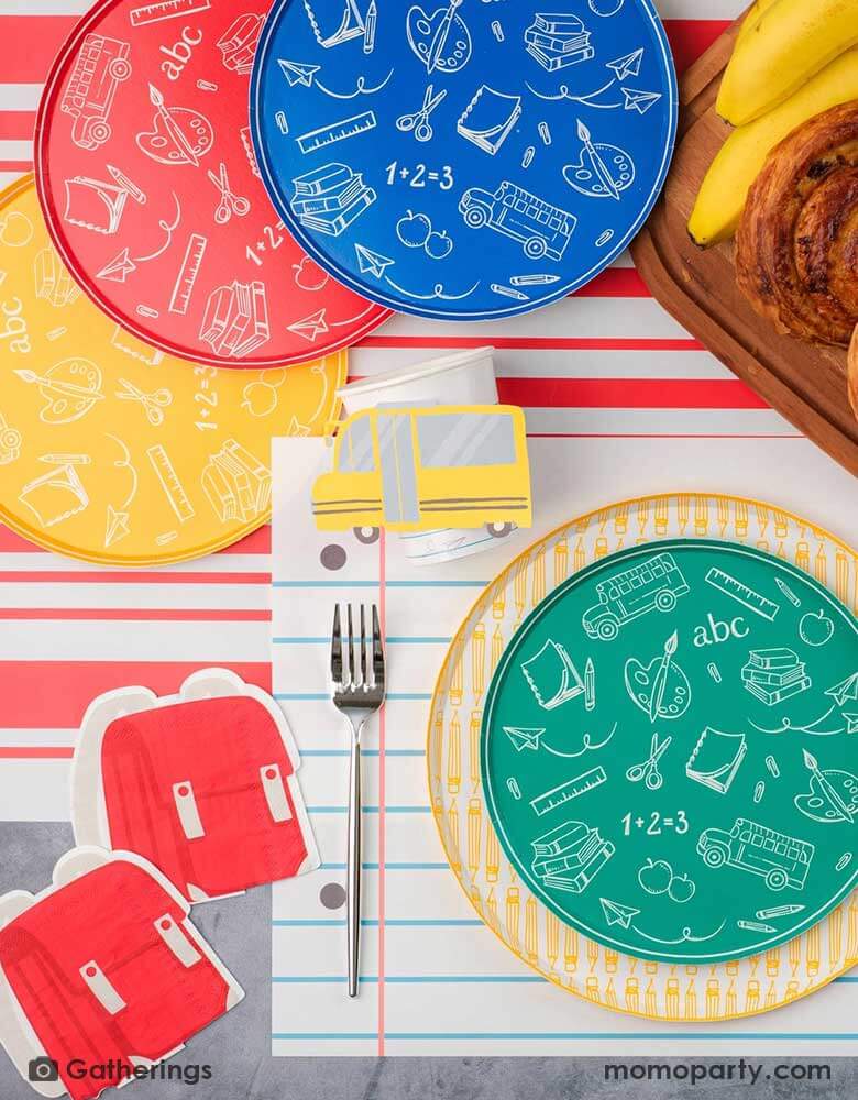 A festive back to school first day of school celebration party table setting featuring Momopartys's back to school themed party supplies including the No. 2 pencil patterned dinner plates, school day side plates with school supplies designs in four different colors, and the notebook inspired placemat by Gatherings. With pencil shaped napkins and red backpack shaped napkins, plus a plate of treats and fruits, this makes a great inspo to celebrate the start of the new school year with kids!