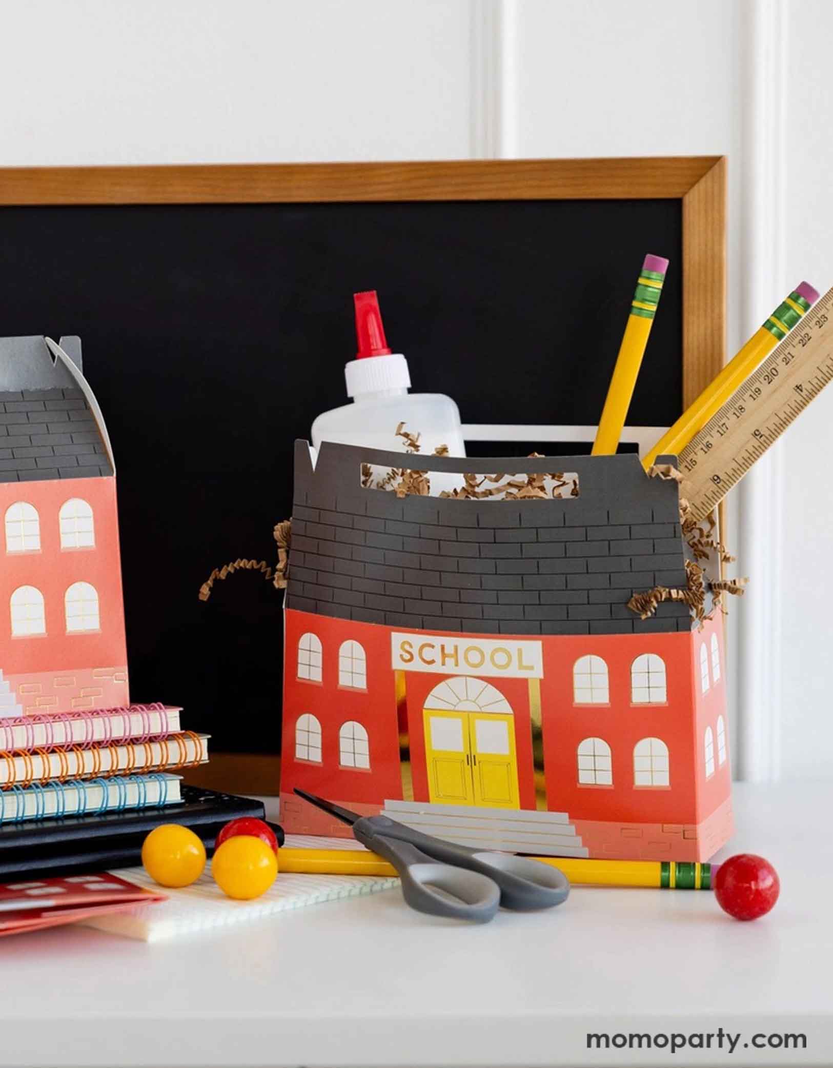 Two of Momopartys's 7" x 2.5" x 5.5" School House Treat Boxes by My Mind's Eye on a desk with school supplies including pencils, rulers, scissors, glue bottles in them. Around the treat boxes there are some colorful notebooks and some red and yellow gum balls and in the back there's a small chalkboard, making this an adorable set up for kid's school themed celebration, be it a back to school party or a first day of school celebration.