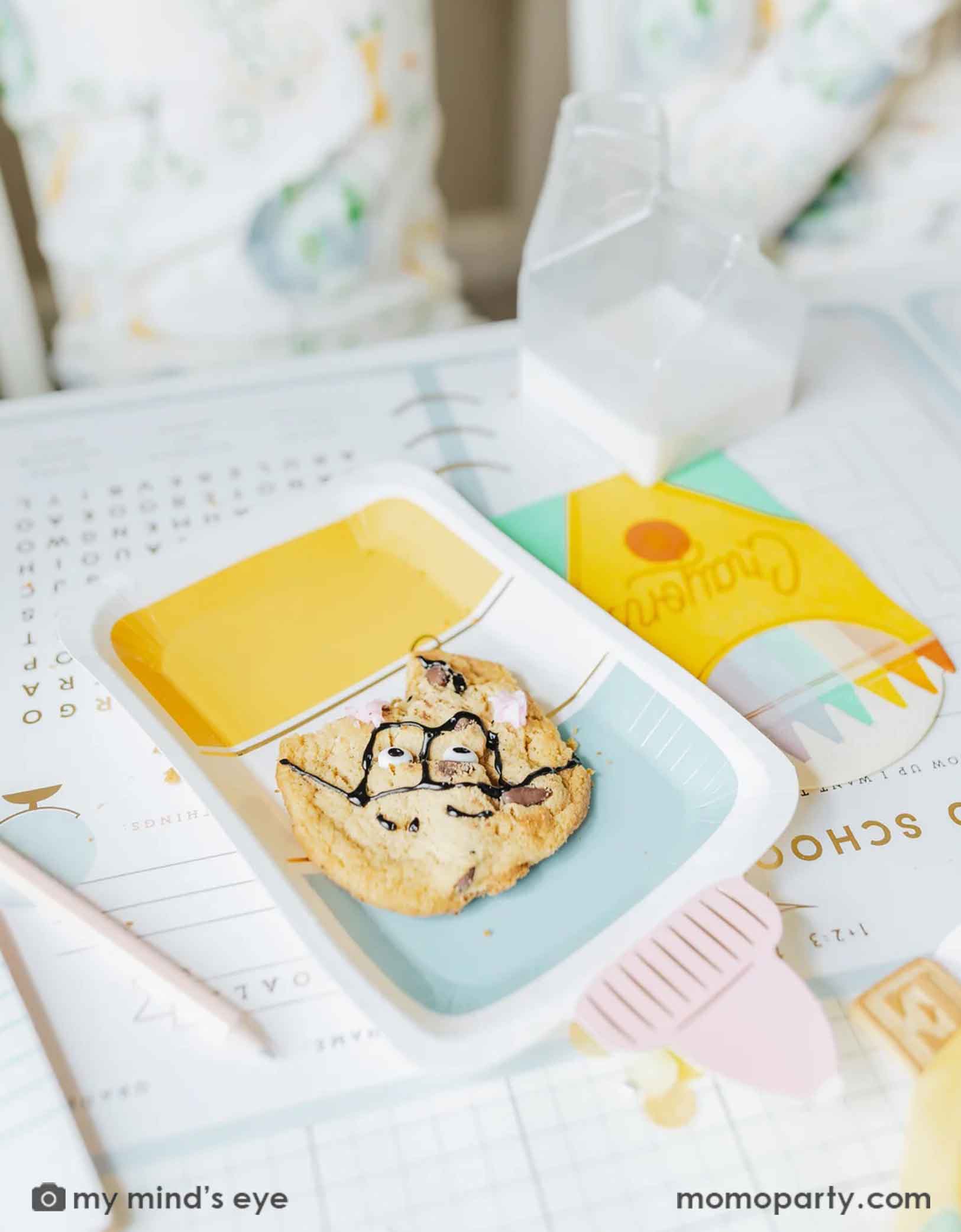 A close up shot of Momopartys's glue bottle shaped party plate by My Mind's Eye with a smart cookie on it. Around the glue bottle plate are some other school themed party supplies including the crayon box shaped napkins, on the back to school activity game placemat, ready for kid's first day of school breakfast celebration!
