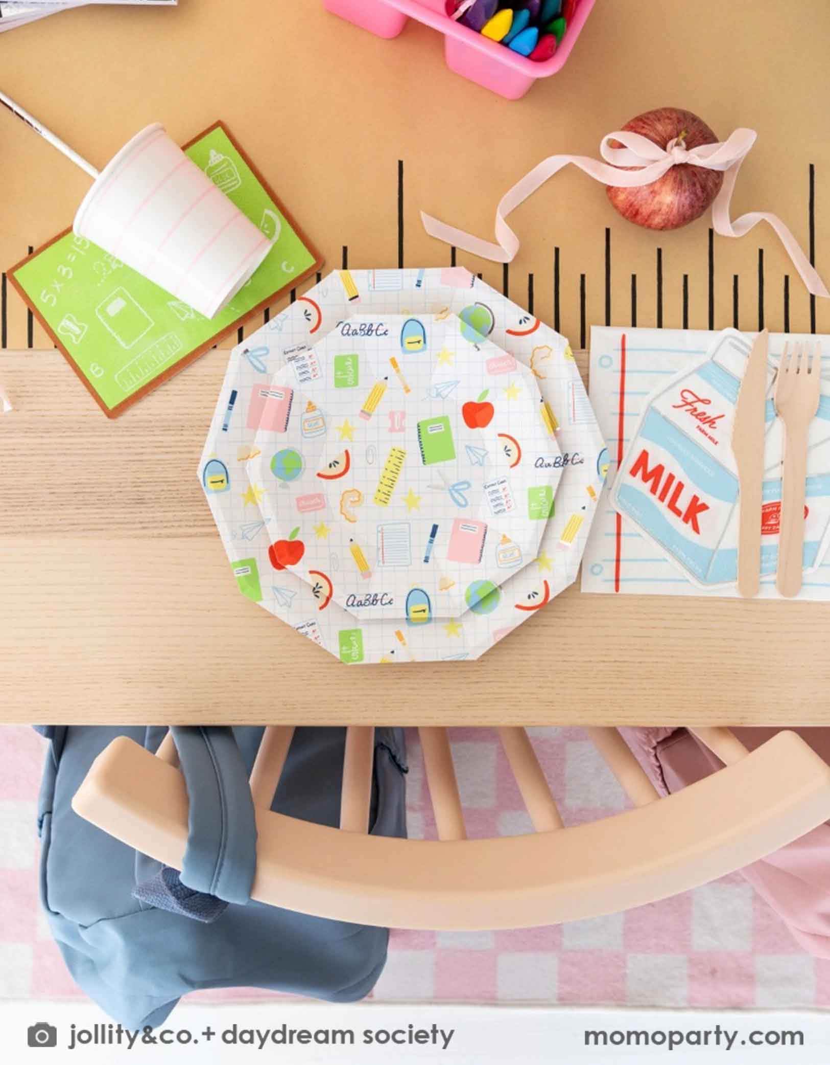 An overhead shot of an adorable kid's school themed party table featuring school supplies themed tablewares from Momopartys including the Smarty Pants milk box shaped napkins, notebook shaped napkins, and pastel pink striped cups, along with school icons patterned plates on a ruler inspired table runner. In the middle of the table there is a pink box with crayons and an apple tied with a pink ribbon, making this an adorable table decor inspiration for kid's first day of school or back to school celebration.
