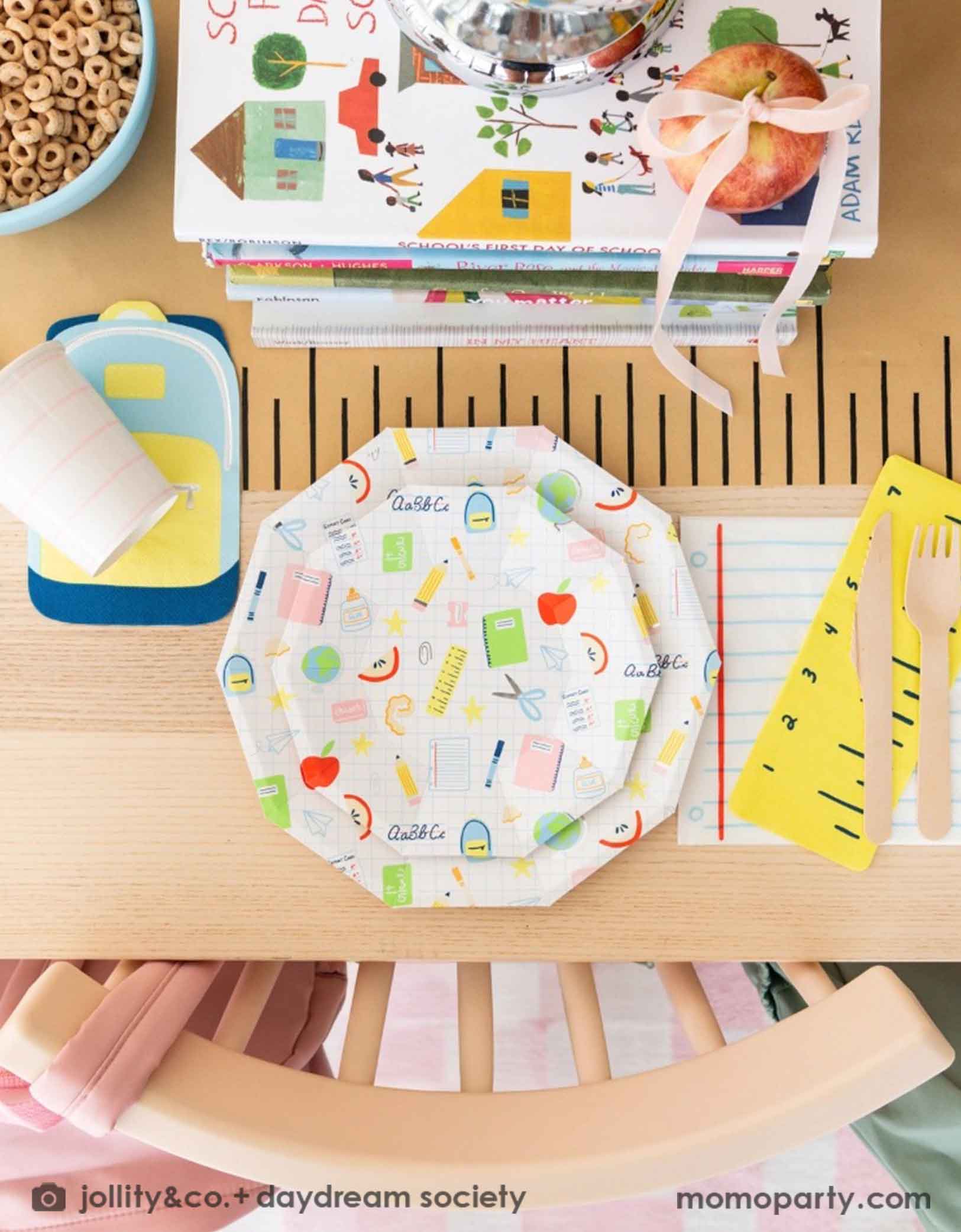 An overhead shot of an adorable kid's school themed party table featuring school supplies themed party tablewares from Momopartys including the Smarty Pants ruler shaped napkins, notebook shaped napkins, and backpack shaped napkins, along with school icons patterned plates, all on a ruler inspired table runner. In the middle of the table there is a pile of books, a bowl of cereal and apples as centerpiece, making this a great decoration inspiration for kid's first day of school or a back to school party.