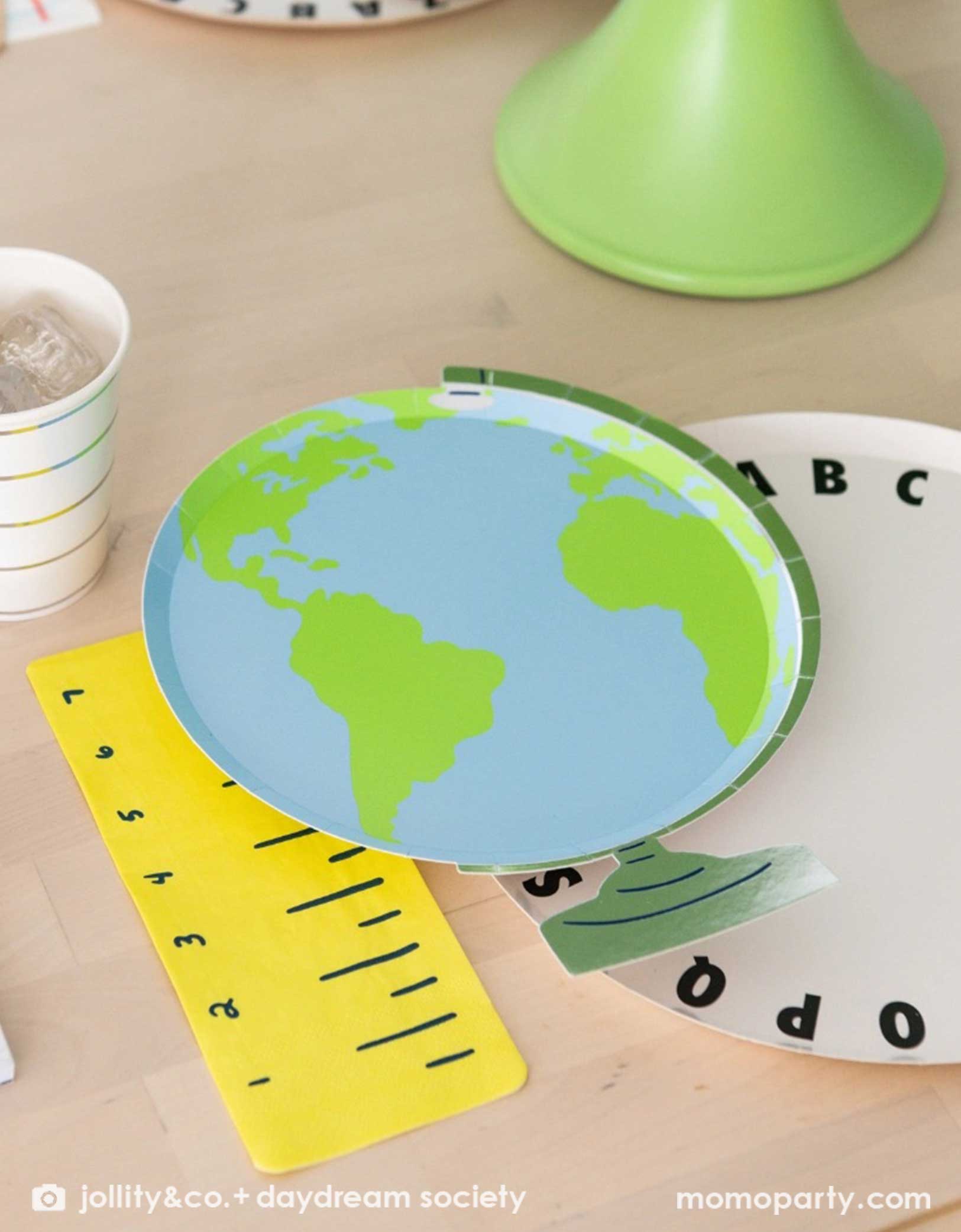 A close up shot of an adorable kid's school themed party featuring school supplies themed party tablewares from Momopartys including the Smarty Pants Globe shaped Large Plate, alphabet round dinner plate, ruler shaped napkin and striped party cup - making this a cute and festive tablescape for kid's school themed celebration, be it the last day of school party, the first day of school celebration or a back to school party.