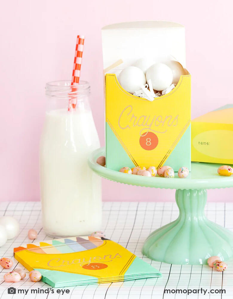 A back to school party table set up in a pink room featuring school themed party supplies from Momopartys including crayon box shaped napkins, crayon box treat boxes by My Mind's Eye on a mint cake stand on a grid table runner next to a milk bottle, with jelly beans around, this makes a great inspiration for a first day of school celebration or kid's back to school parties.