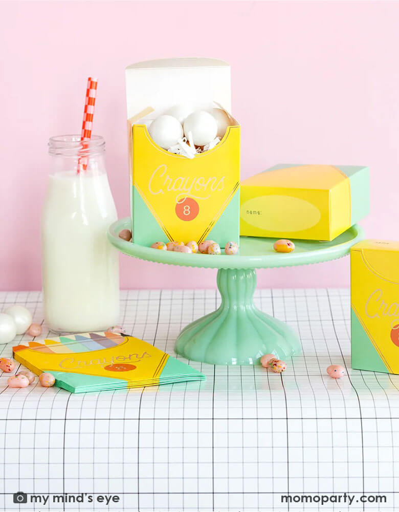 A back to school party table set up in a pink room featuring school themed party supplies from Momopartys including crayon box shaped napkins, crayon box treat boxes by My Mind's Eye on a mint cake stand on a grid table runner next to a milk bottle, with jelly beans around, this makes a great inspiration for a first day of school celebration or kid's back to school parties.