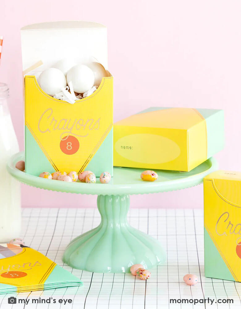 A back to school party table set up in a pink room featuring school themed party supplies from Momopartys including crayon box shaped napkins, crayon box treat boxes by My Mind's Eye on a mint cake stand on a grid table runner next to a milk bottle, with jelly beans around, this makes a great inspiration for a first day of school celebration or kid's back to school parties.