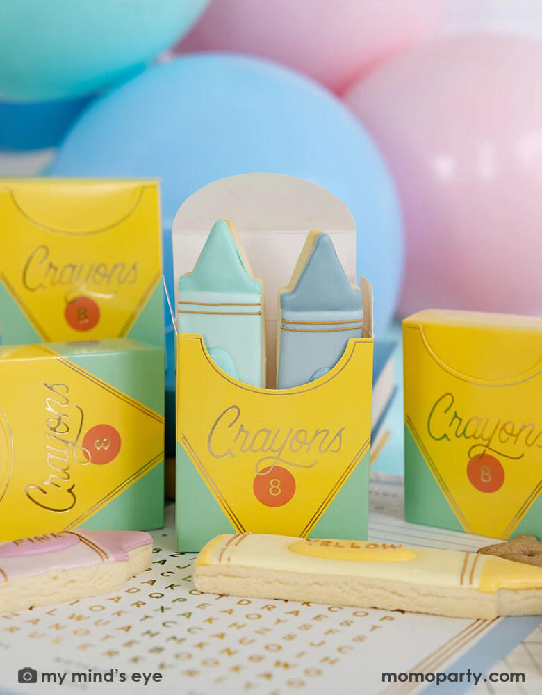 A tableshot of a back to school party featuring crayon shaped treat boxes from Momopartys. The treat boxes are filled with crayon shaped sugar cookies in adorable pastel colors, in the back there are some pastel colored party balloons, making this an adorable idea for kid's first day of school celebration party.