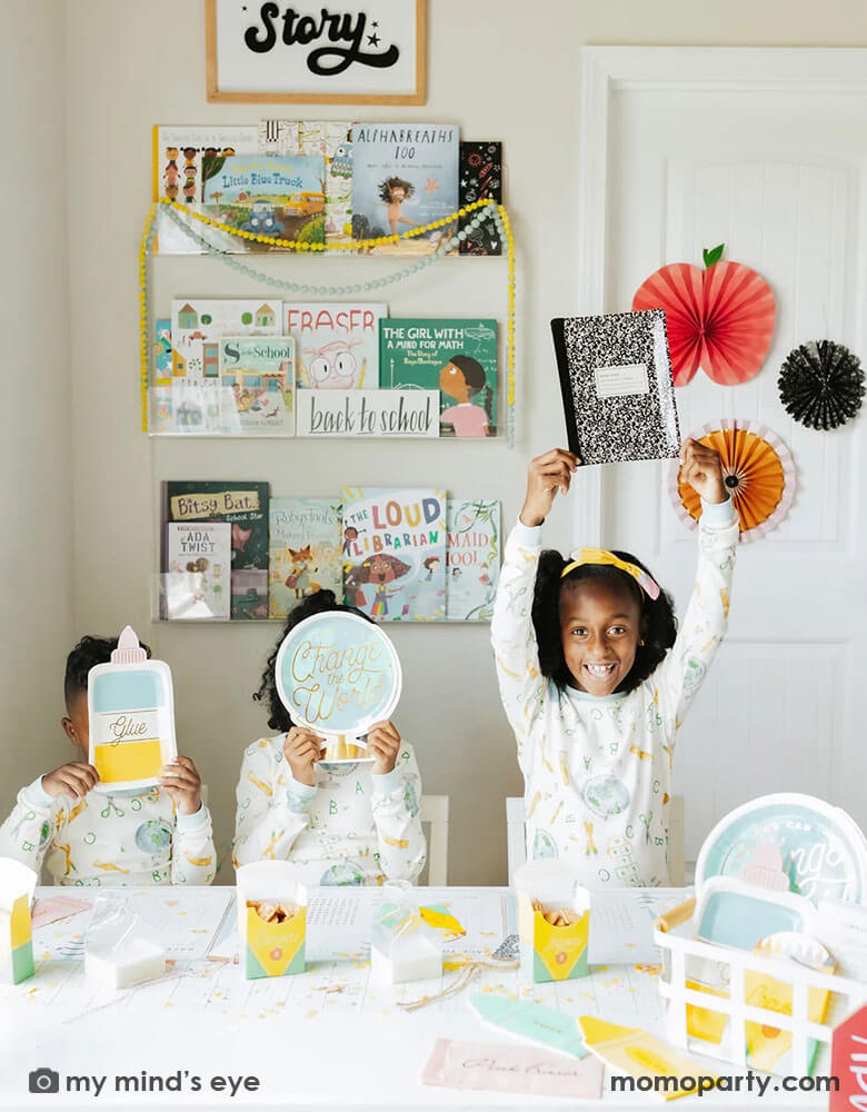 A first day of school breakfast party with three siblings in their school themed pajamas at their house. The kids are holding the notebook shaped plate, the globe shaped plate, the glue bottle shaped plate. On the table there are more school themed party supplies from Momopartys including crayon shaped treat boxes, eraser shaped napkins, notebook shaped plates. On the wall there are also some apple and pencil shaped party fans as the backdrop. A simple yet cute way to celebrate learning!