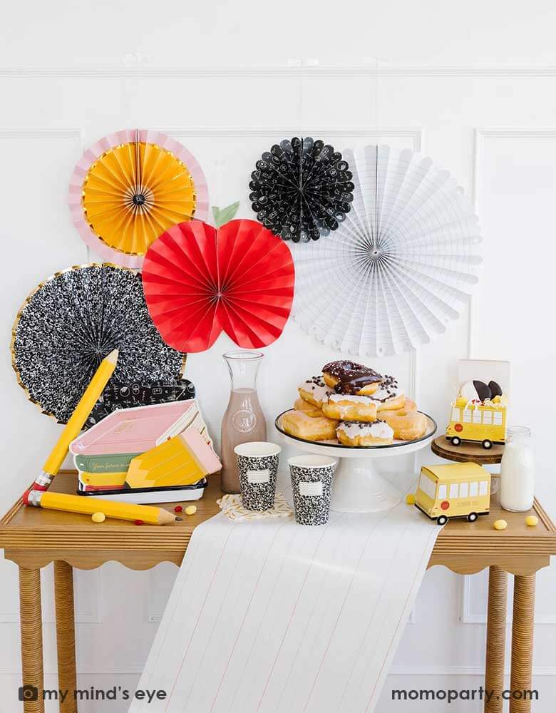 A school themed party table featuring school themed party supplies from Momopartys including stack of books plates, pencil shaped napkins, composition notebook party cups, school bus treat boxes and writing paper table runner around some donuts and milk bottles. The back wall is adorned with back to school party fans by My Mind's Eye including an apple, an pencil, a notebook, an equation pattern and a composition notebook fan. A perfect inspo for kid's back to school, first day of school celebration.