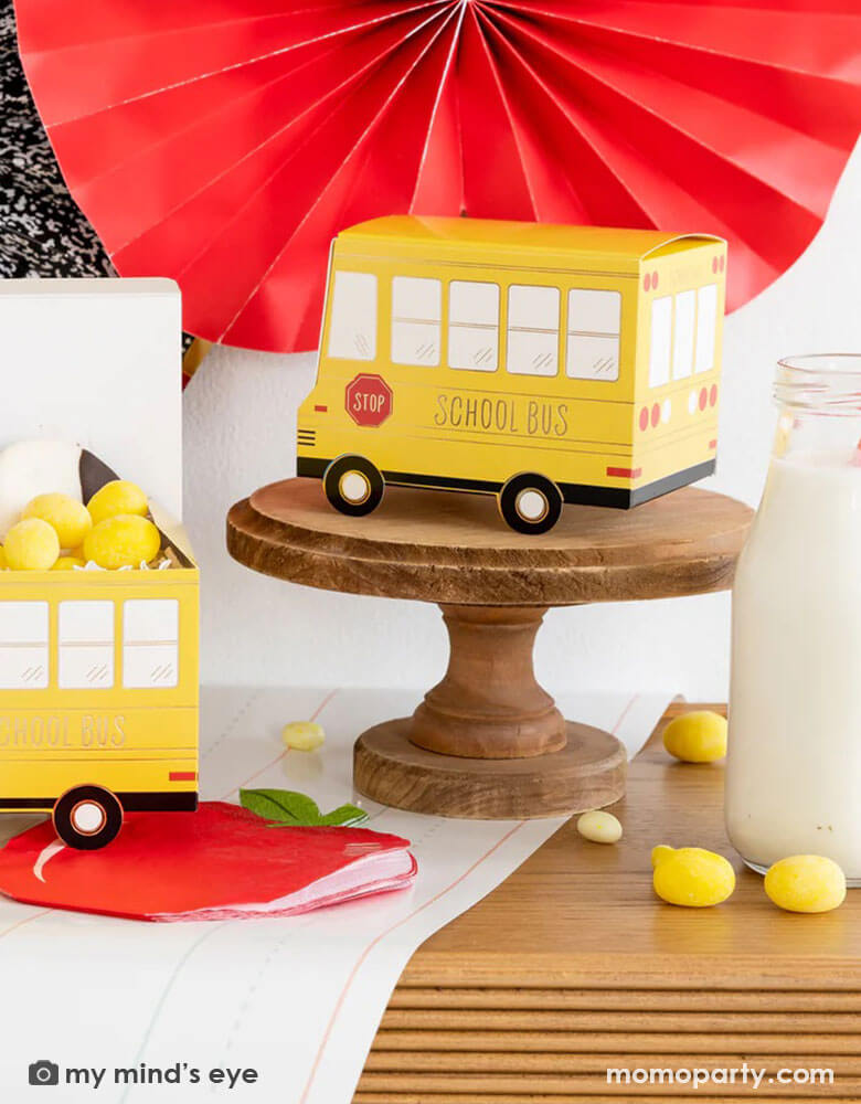 A festive back to school party table features Momopartys's school themed party supplies including school bus shaped treat boxes, apple shaped napkins, writing paper table runner by My Mind's Eye. On the table are some red jelly beans, yellow candies and milk bottles in coordinating colors. In the back there's the back to school party fan hung on the wall, making this a perfect inspiration for kid's back to school party or a first day of school celebration breakfast.
