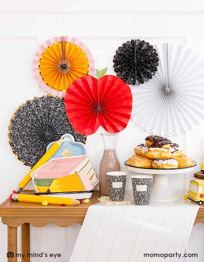 A party table featuring school themed party supplies from Momopartys including stack of books plates, backpack plates, pencil shaped napkins, composition notebook party cups, school bus treat boxes and writing paper table runner around some donuts and milk bottles. The back wall is adorned with back to school party fans by My Mind's Eye including an apple, an pencil, a notebook, an equation pattern and a composition notebook fan. A perfect inspo for kid's back to school, first day of school celebration.