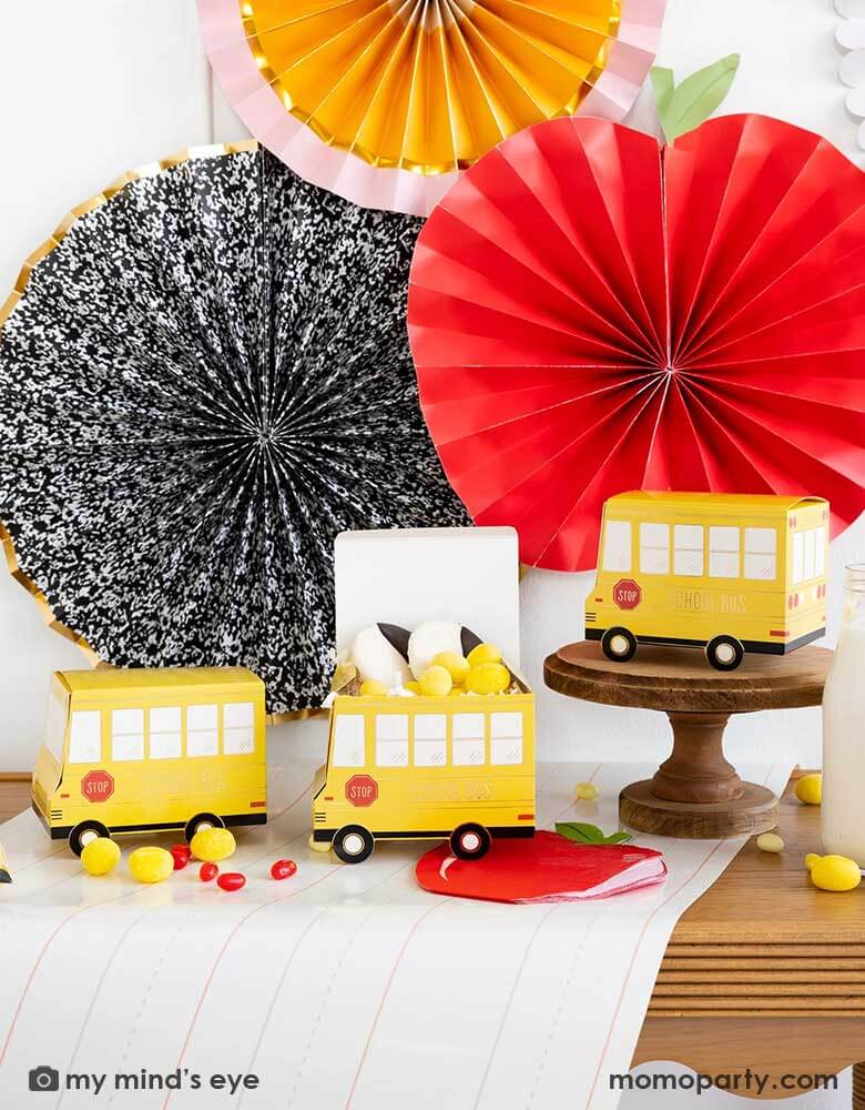 A festive back to school party table features Momopartys's school themed party supplies including school bus shaped treat boxes, apple shaped napkins, writing paper table runner by My Mind's Eye. On the table are some red jelly beans, yellow candies and milk bottles in coordinating colors. In the back there's the back to school party fan hung on the wall, making this a perfect inspiration for kid's back to school party or a first day of school celebration breakfast.