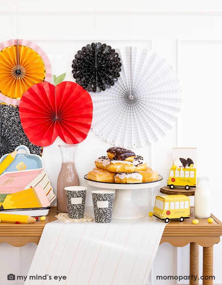 A party table featuring school themed party supplies from Momopartys including stack of books plates, backpack plates, pencil shaped napkins, composition notebook party cups, school bus treat boxes and writing paper table runner around some donuts and milk bottles. The back wall is adorned with back to school party fans by My Mind's Eye including an apple, an pencil, a notebook, an equation pattern and a composition notebook fan. A perfect inspo for kid's back to school, first day of school celebration.