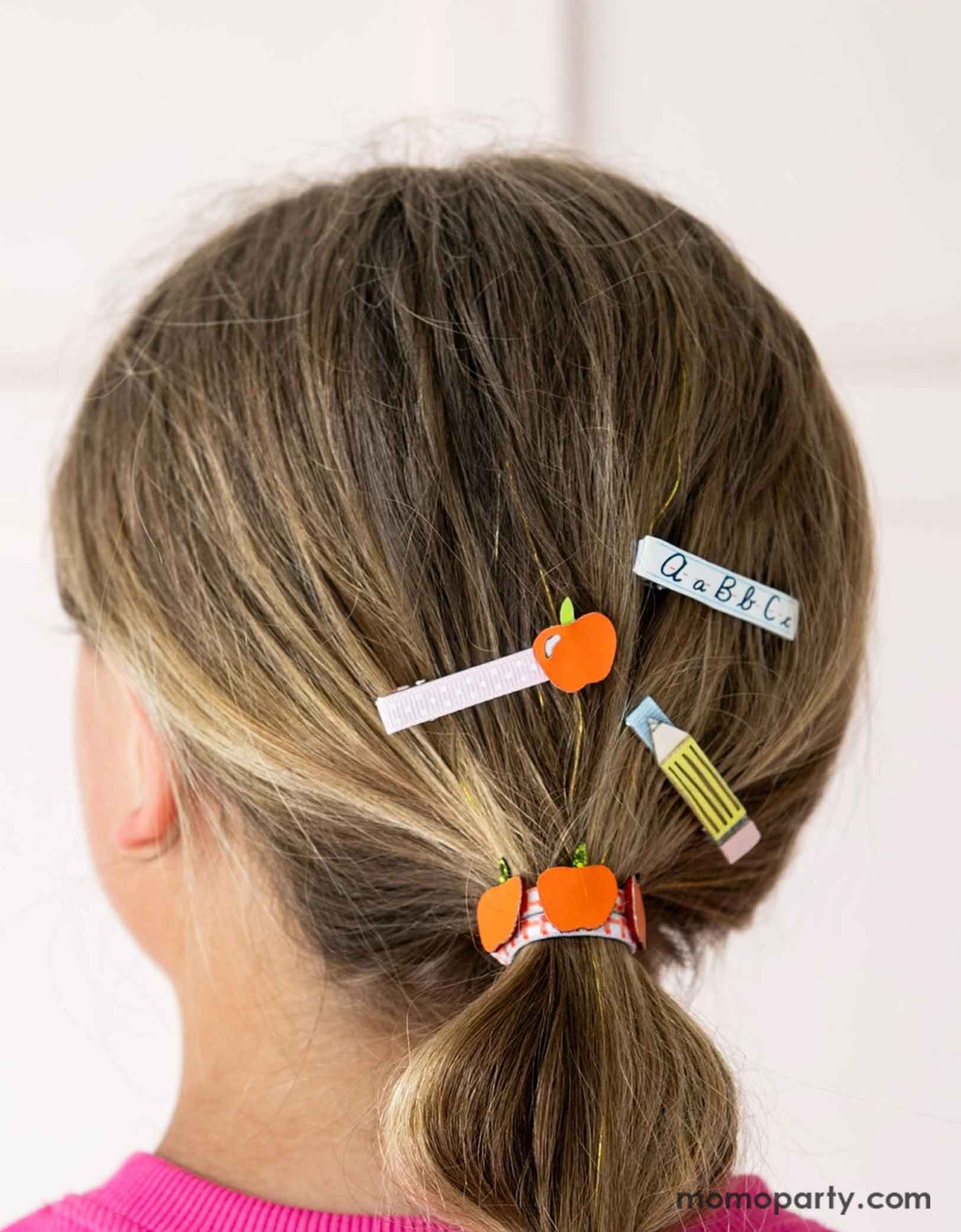 A girl with hair styled in a ponytail using Momopartys's Smarty Pants ABC Hair Bar Clip Set by Daydream Society. Three hair clips, one with alphabet letters A, B, and C, one with pencil design, and one with an apple design. These hair clips are perfect for back to school picture day, first day of school, and every school day in between.