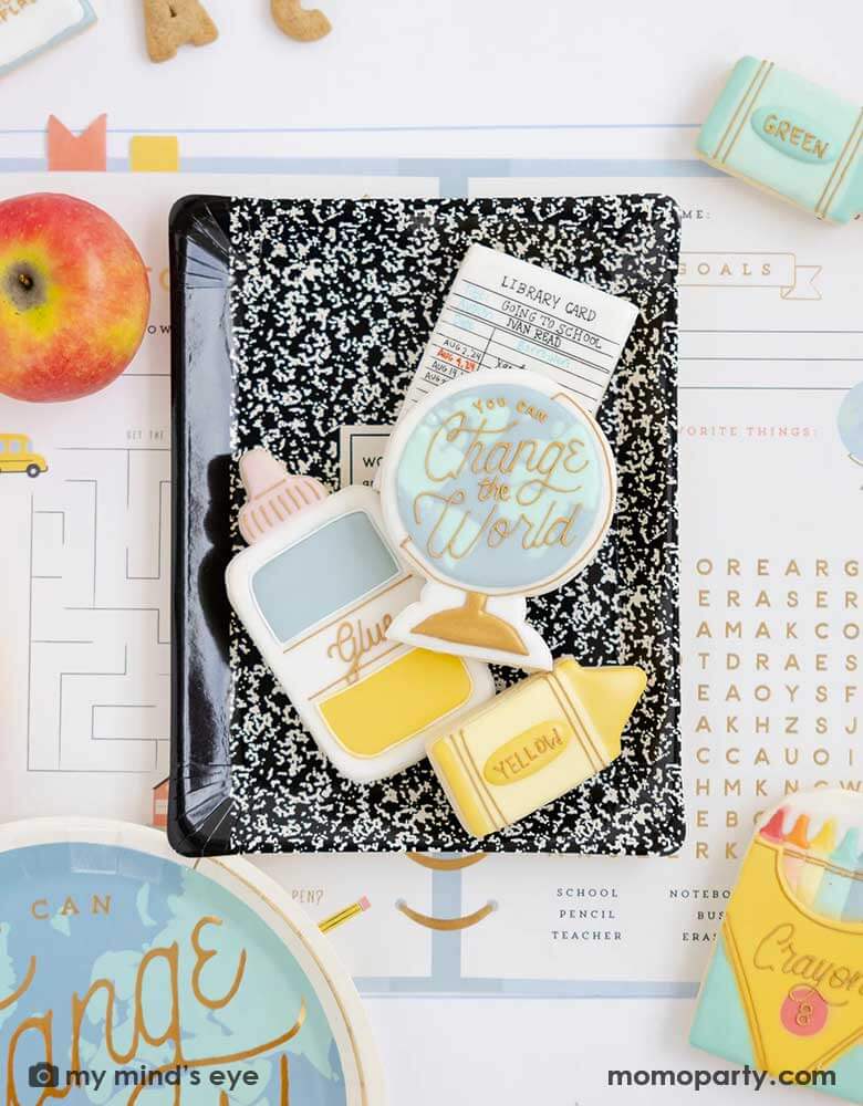 A overhead shot of a place setting from a back to school party featuring Momopartys's school themed party supplies including the back to school planner placemats, a black notebook shaped plate which has multiple matching school supplies themed sugar cookies including crayons, a glue bottle, a globe, a library card on it, with an apple and some alphabet shaped cookies over the placemat, this makes a festive and adorable table setting ideas for kid's first day of school celebration.