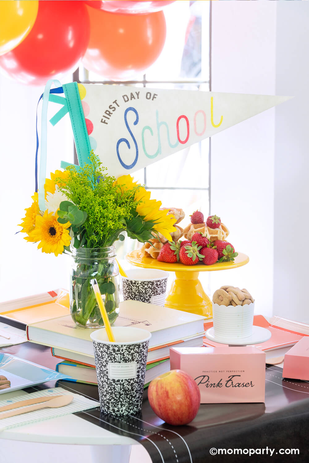 An adorable first day of school breakfast table setting featuring school themed party goods from Momopartys including the first day of school felt party pennant, chalkboard table runner, pink eraser shaped treat boxes, composition notebook party cups with pencil straws, and text book shaped plates. There are also some waffles stacked and strawberries on a cake stand and a sunflower bouquet. With festive balloons in the back, it's a perfect set up for your child's first day of school celebration.