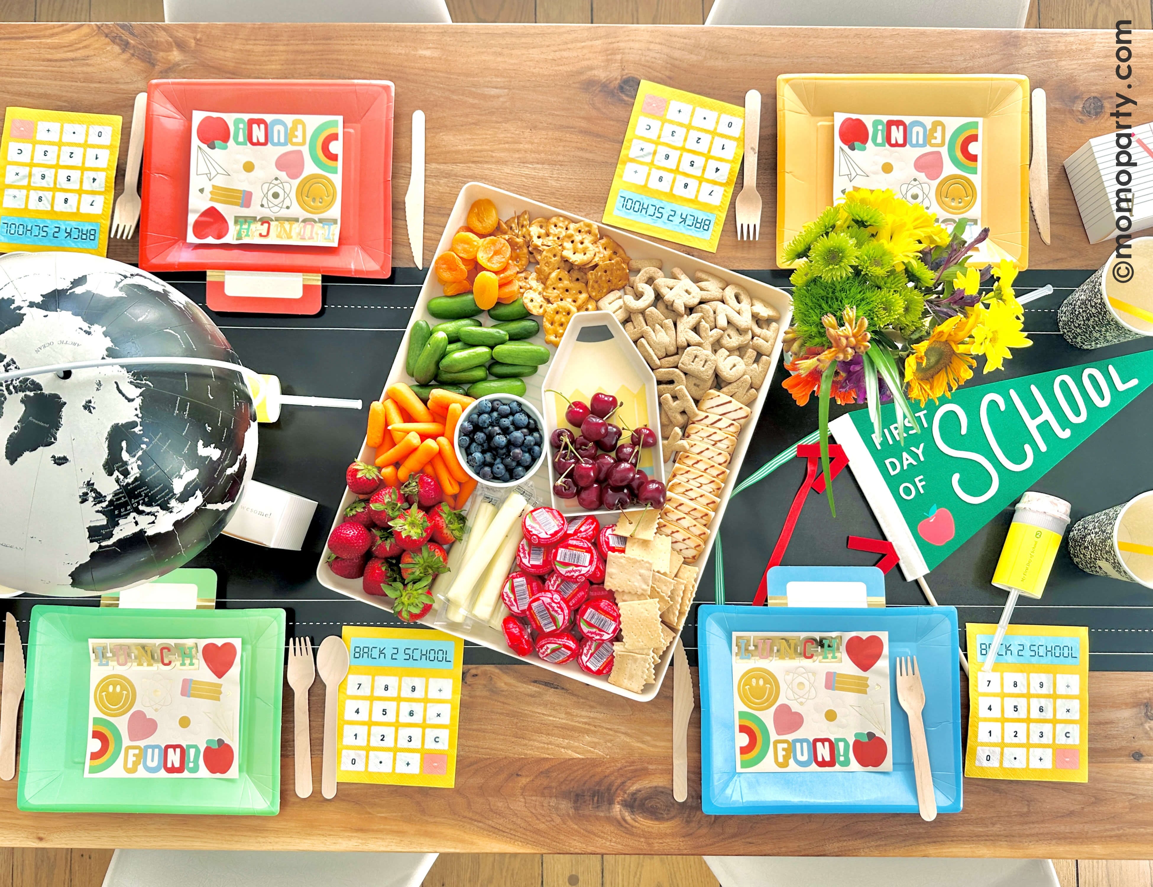 A overhead shot of a festive back to school party table set up featuring various school themed party decors and tableware from Momopartys including the chalkboard table runner, lunchbox shaped party plates, calculator shaped napkins, notebook party cups, the first day of school apple felt party pennant, and pencil shaped confetti poppers. In the middle of the table is the paper and pencil bamboo tray set which is filled with healthy snacks for a fun back to school playdate to celebrate the new school year!