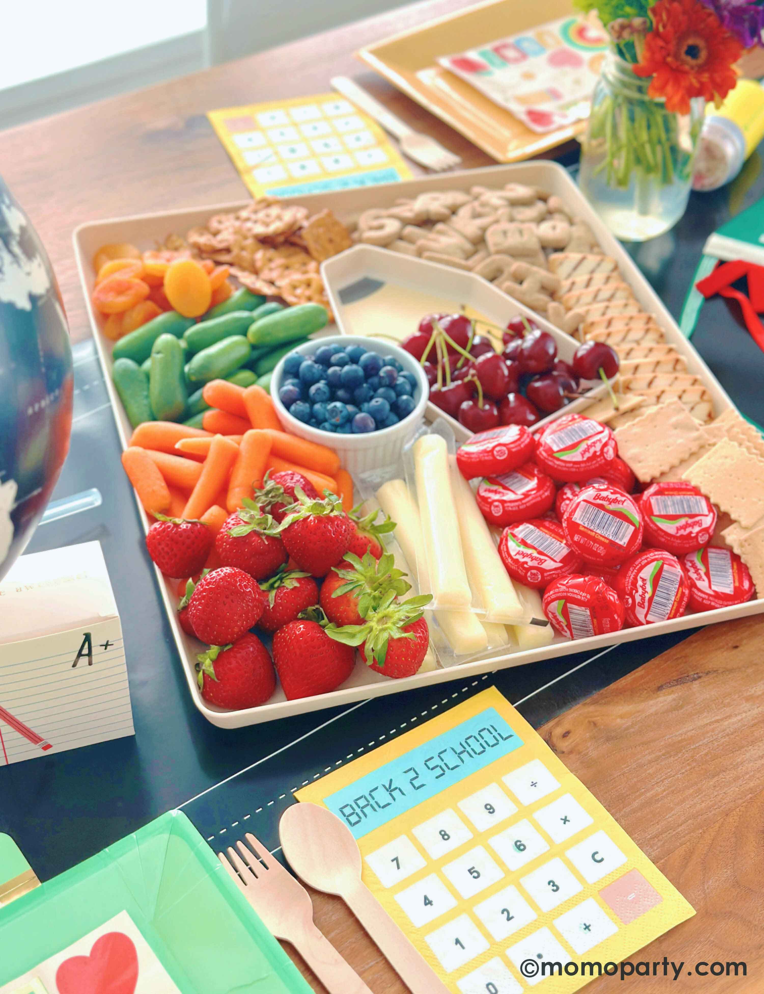 A close-up shot of the paper and pencil bamboo tray set from Momopartys which is filled with healthy snacks and treats including fruits, vegetables, cheeses, pretzels and cookies at a kid's back to school party. The tray is surrounded by multiple school themed party tablewares including notebook shaped treat boxes, calculator shaped napkins, and lunchbox shaped plates. This gives a great idea for a fun back to school playdate for your child and their friends to celebrate the new school year!