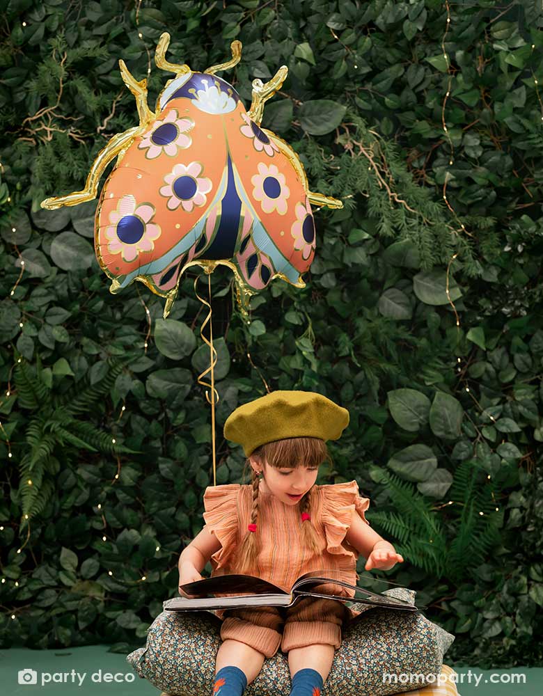 A little girl reading on her book in the backyard with a wall of greenery, next to her is Momopartys's 34 x 30 inches ladybird shaped foil balloon by Party Deco. With coral color with small flower pattern with gold legs on it.
