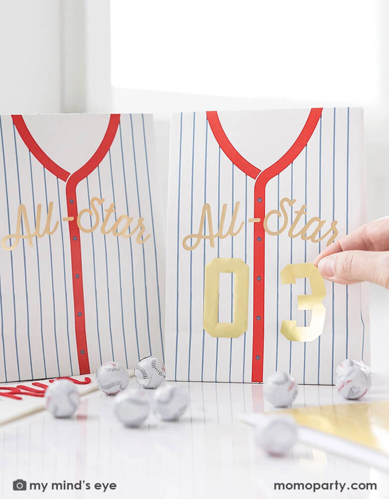 A baseball party table features Momopartys's baseball treat bags by My Mind's Eye. A hand is putting the gold number stickers to the bad to personalize the favor bag. On the table there are some baseball chocolate balls. With these treat bags, they make great party favor for your kid's friends at their baseball themed party!