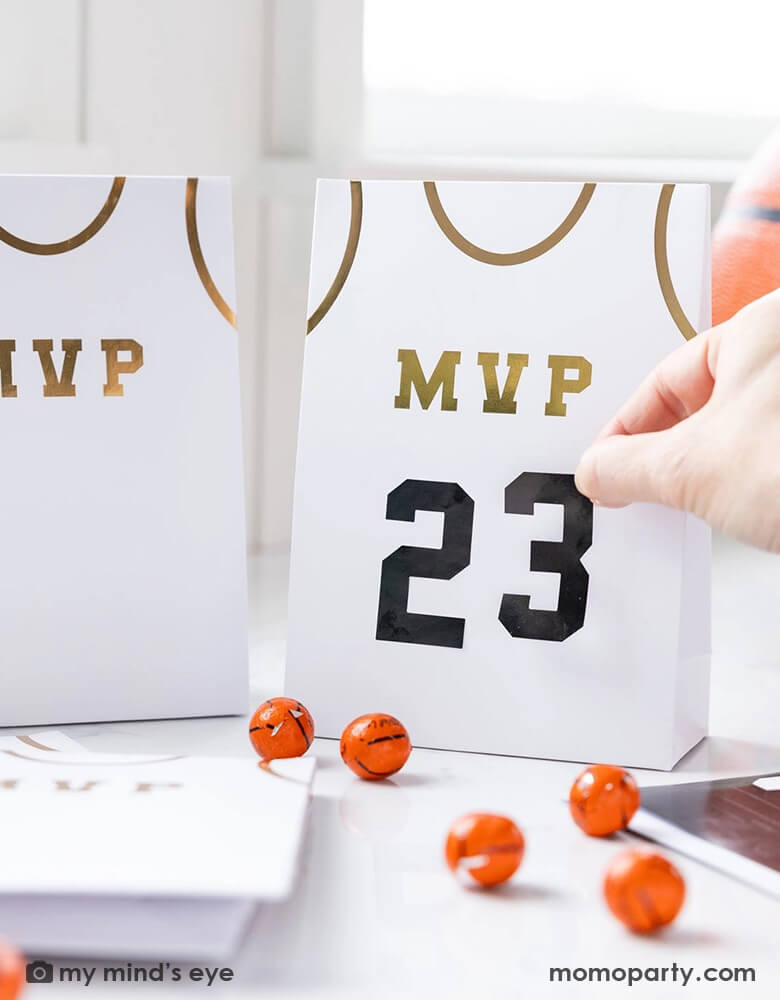 A basketball themed party table features Momopartys's basketball treat bags by My Mind's Eye. Comes in a set of 8 treat bags, these basketball jersey shaped treat bags with MVP on them in gold foil are personalizable with the black number stickers. On the table there are some basketball chocolate balls, makes this a perfect idea for party favors at kid's basketball themed birthday party. 
