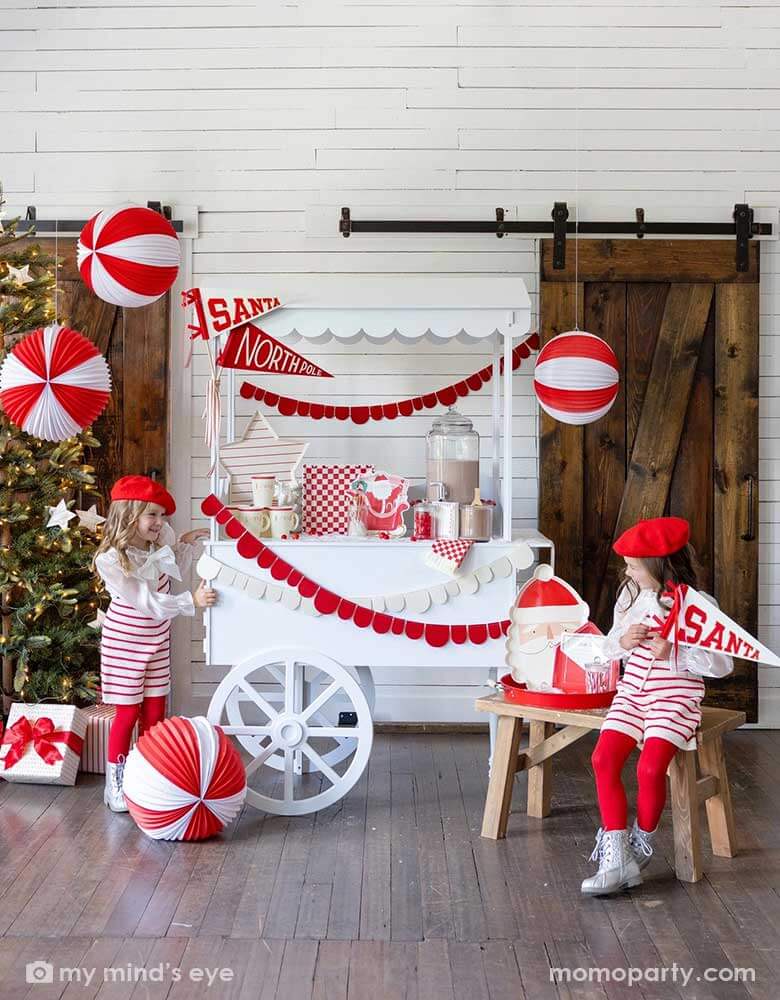 Two girls in an adorable Santa themed hot cocoa station set up for a kid's Holiday party featuring a white dessert cart with hot chocolate and Momopartys's Dear Santa party supplies including Santa shaped party cups, checkered plates, napkins, Santa on a sleigh shaped plates and Dear Santa envelope shaped plates. Around the hot chocolate station are two felt party pennant with Santa & Northpole written on them, red scalloped shaped party garlands and some red and white honeycomb balls hung from the ceiling.