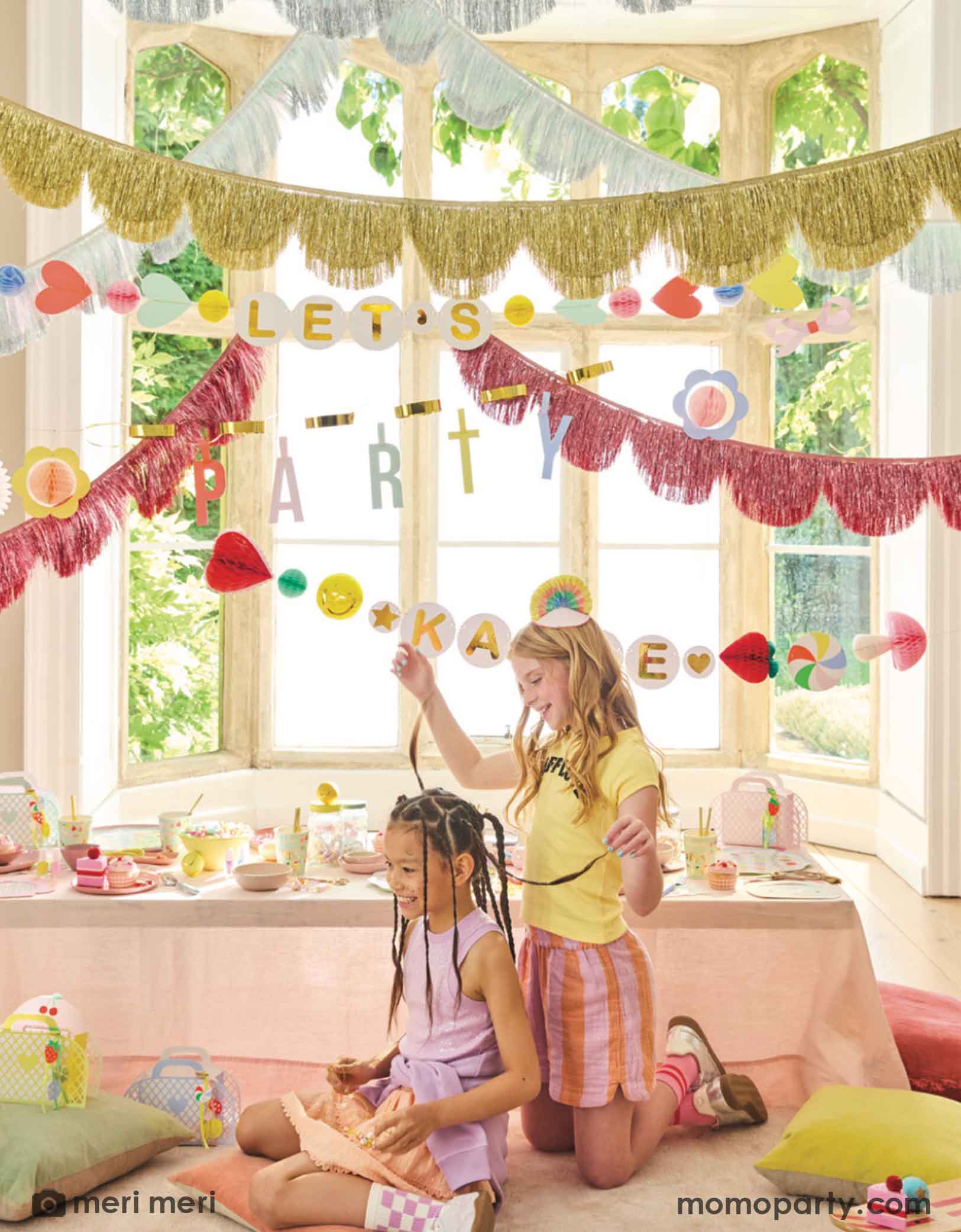 Two pre-teen girls in a room decorated with colorful Momopartys's Charm Party Collection by Meri Meri including festive banners, metallic fringe garlands, and a table of charm party themed tablewares including plates, napkins and cups. A perfect set up for girl's sleepover party or a BFF gathering.
