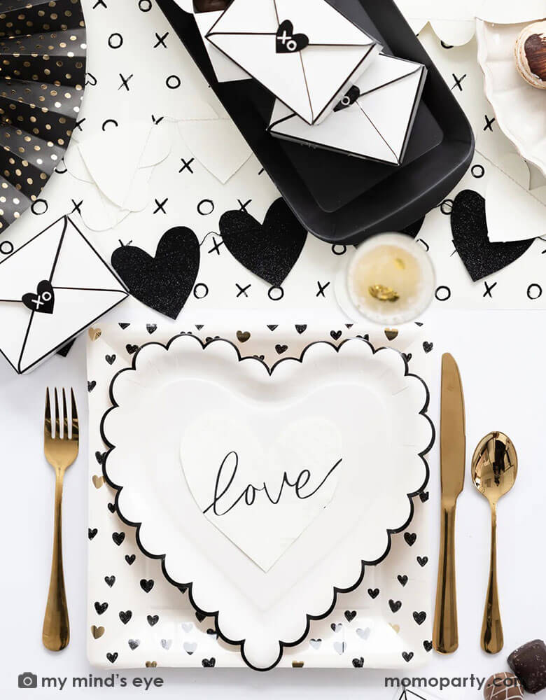 A black and white sophisticated Valentine's Day party tablescape featuring Momopartys's Valentine's Day party supplies including the XOXO paper table runner, heart shaped plates and black heart scattered dinner plates.