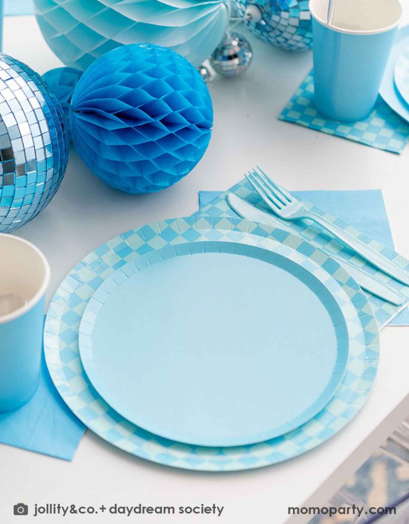 A blue themed party table features Momopartys's Blue and Check it! Blue Checkered Tableware Collection by Jollity & Co. including 10" blue checkered dinner plates, 8" blue dessert plates, 6.5" blue checkered large napkins, 5" blue small napkins, blue plastic utensils and solid blue party cups, with blue honeycomb and disco ball decorations as the centerpiece for the party table.