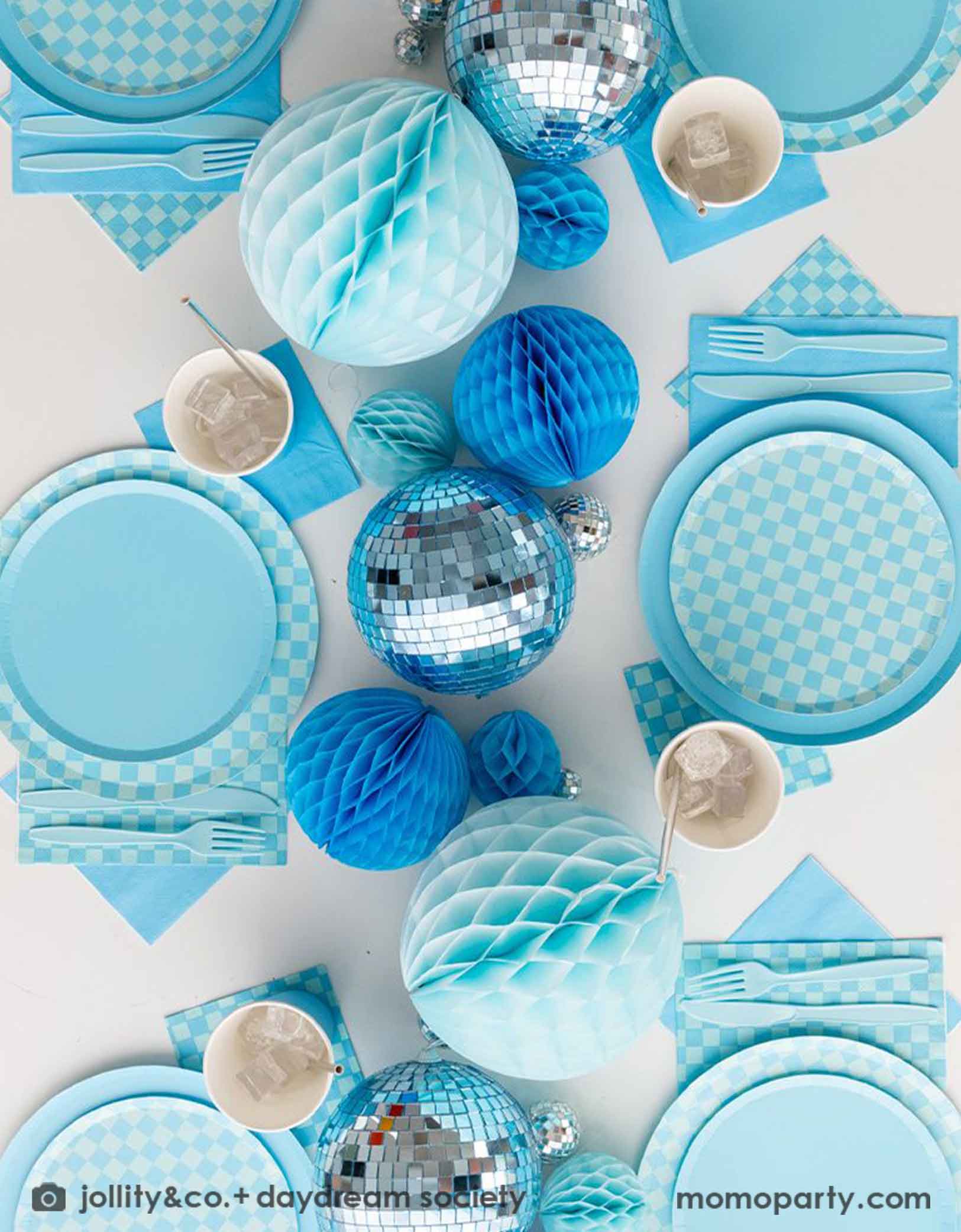 A overhead shot of a blue themed party table features Momopartys's Blue and Check it! Blue Checkered Tableware Collection by Jollity & Co. including 10" blue dinner plates, 8" blue checkered dessert plates, 6.5" large napkins, blue plastic utensils and solid blue party cups, with blue honeycomb and disco ball decorations as the centerpiece for the party table.