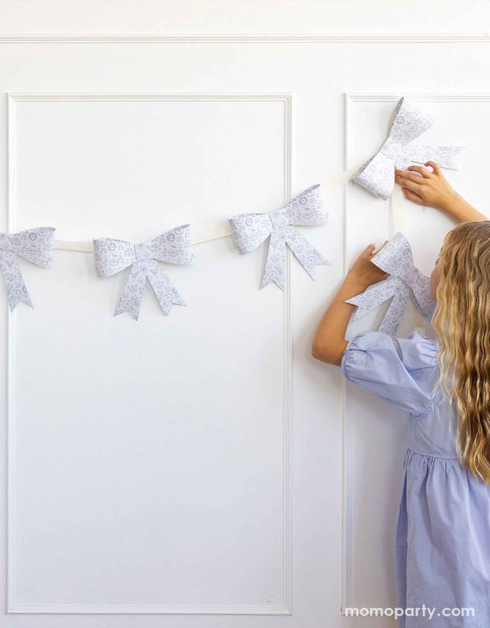 A girl in pastel blue dress holding and hanging Momopartys's 7' Floral Bow Banner onto a white wall. Made of high-quality paper, this garland features delicate flowers and a cute bow design. Perfect for weddings, baby showers, or garden parties. Spring into style with this beautiful banner!