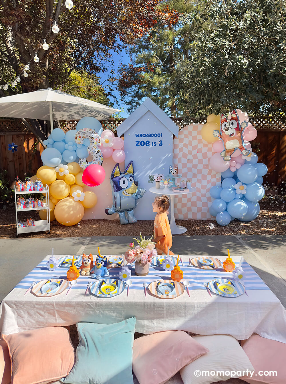 A little girl playing Keepy Uppy balloon game at a Bluey themed party set up by Momopartys featuring pastel color balloon garlands, Bluey shaped foil balloons, a kid's picnic table with Bluey inspired tablewares, decorations and pastel pillows around it.