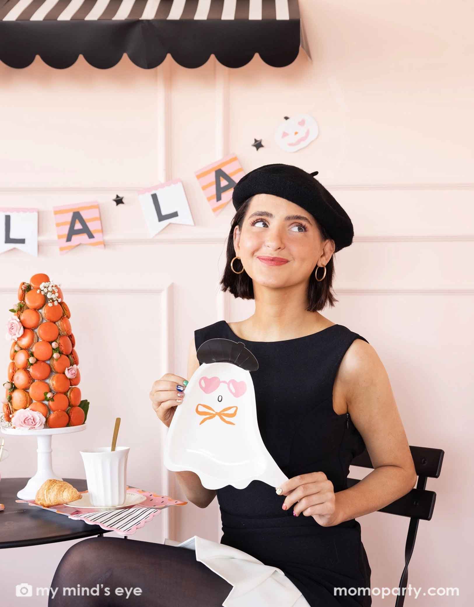 A lady in black dress and beret holding Momopartys's Boo La La french inspired ghost-shaped plate at a Halloween-themed party decorated with french-inspired party supplies including striped Boo La La party banner, striped napkins, and decorations, , along with a macaron tower on the table.