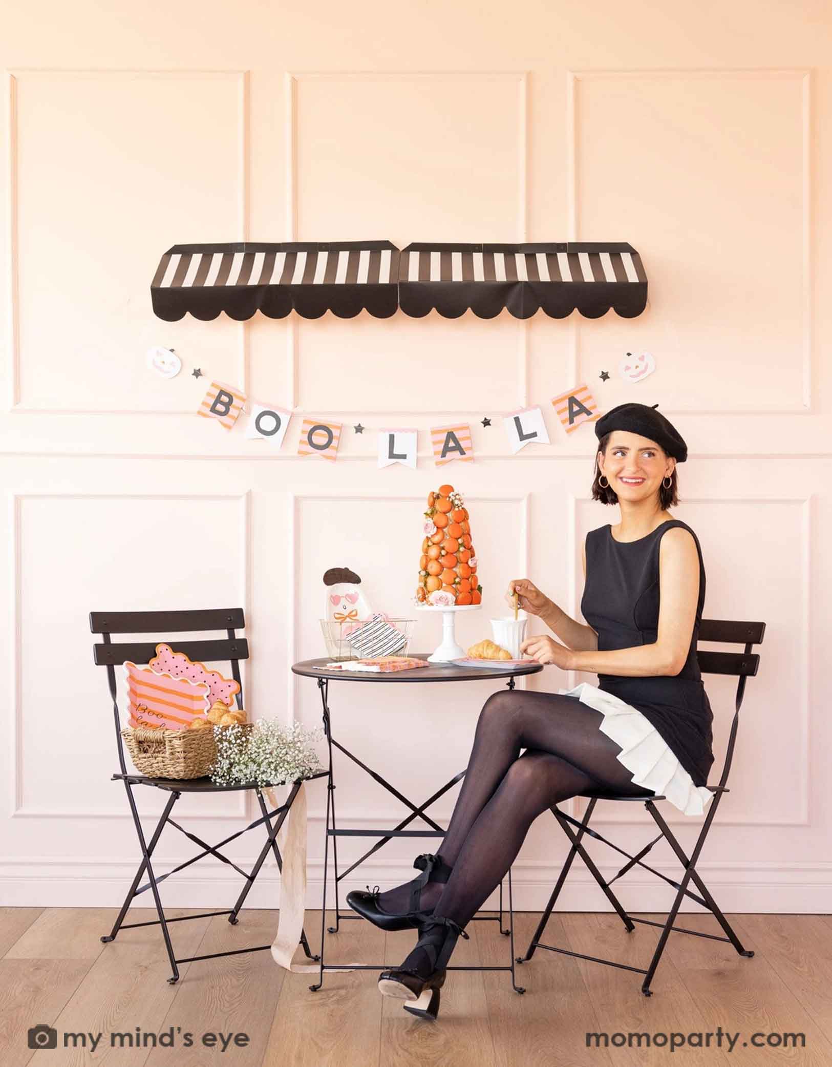 A lady in black dress and beret sitting in a French inspired pink Halloween set up featuring Momopartys's Boo La La party collection which includes ghost-shaped plates, black striped with pink edge napkins, orange and pink striped plates and in a basket with croissants and flowers and a macaron tower on the table. In the background there's a black striped awning and underneath hung the Boo La La striped party banner. A chic idea for a not-so-spooky ghost themed Halloween celebration for girls gathering.