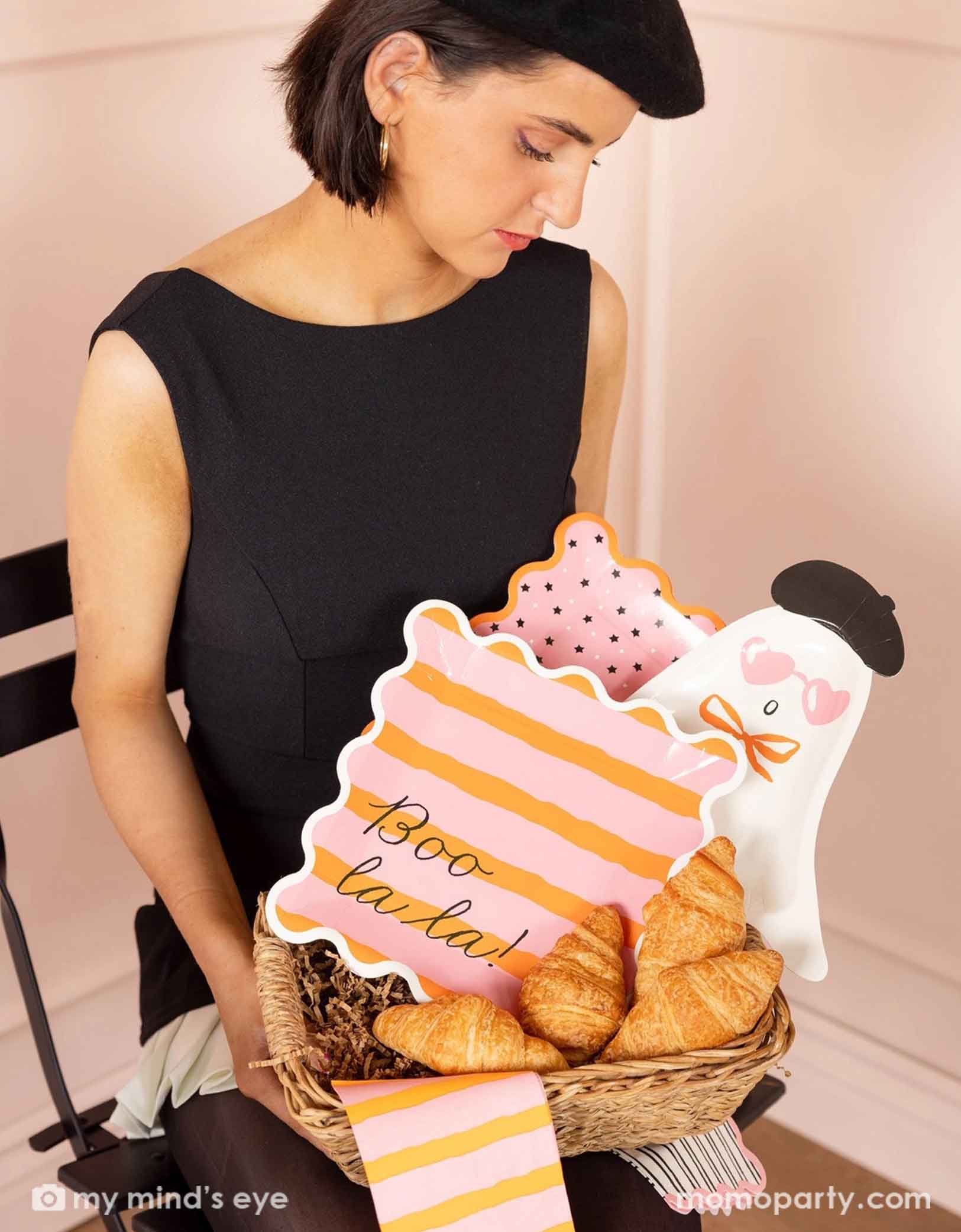 A lady in a black dress and a beret hat holding a french inspired basket filled with Momopartys's Boo La La party tableware collection including the ghost shaped plates, the orange and pink striped dinner plates & napkins, and the pink and black star plates, with croissants around the tablewares.