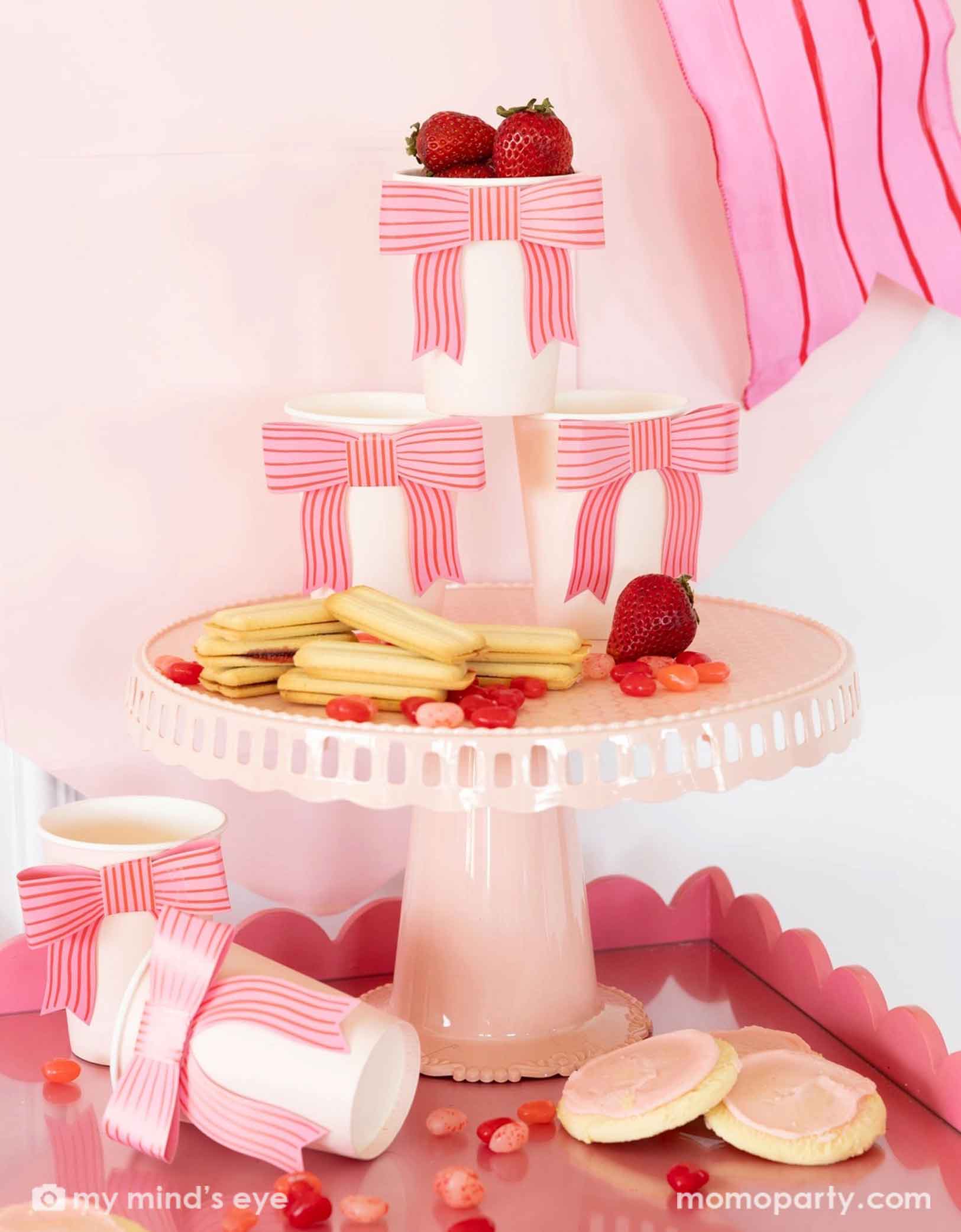 A gorgeous Valentine's Day or Galentine's Day party set up featuring Momopartys's bow party cups staggered on a pink cake stand. On the tray and the stand there are pink and red themed desserts / snacks including sugar cookies, strawberries, jelly beans and more. Making this a great inspiration for a darling Vday celebration set up!