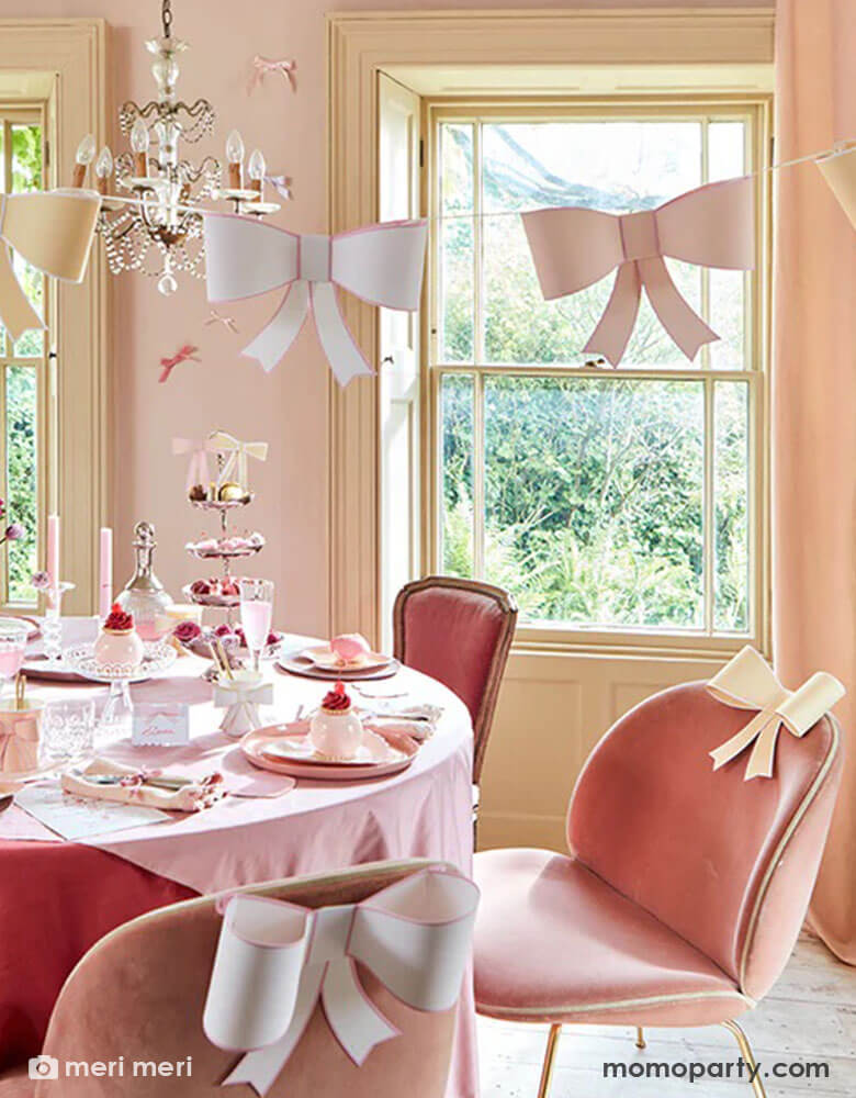 An elegant and chic bow themed party set up featuring Momopartys's pastel heart shaped plates and napkins and 3D bow party cups on the table, above the table there's an elegant chandelier with Meri Meri's 3D bow garland hung over. On the table there are scrumptious treats and sweets for tea time. The chairs are also adorn with 3D bow decoration for a playful touch.