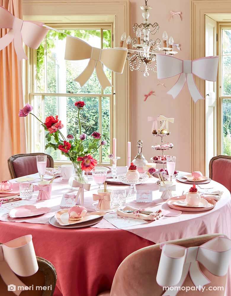 An elegant and chic bow themed party set up featuring Momopartys's pastel heart shaped plates and napkins and 3D bow party cups on the table, above the table there's an elegant chandelier with Meri Meri's 3D bow garland hung over. On the table there are scrumptious treats and sweets for tea time. The chairs are also adorn with 3D bow decoration for a playful touch.